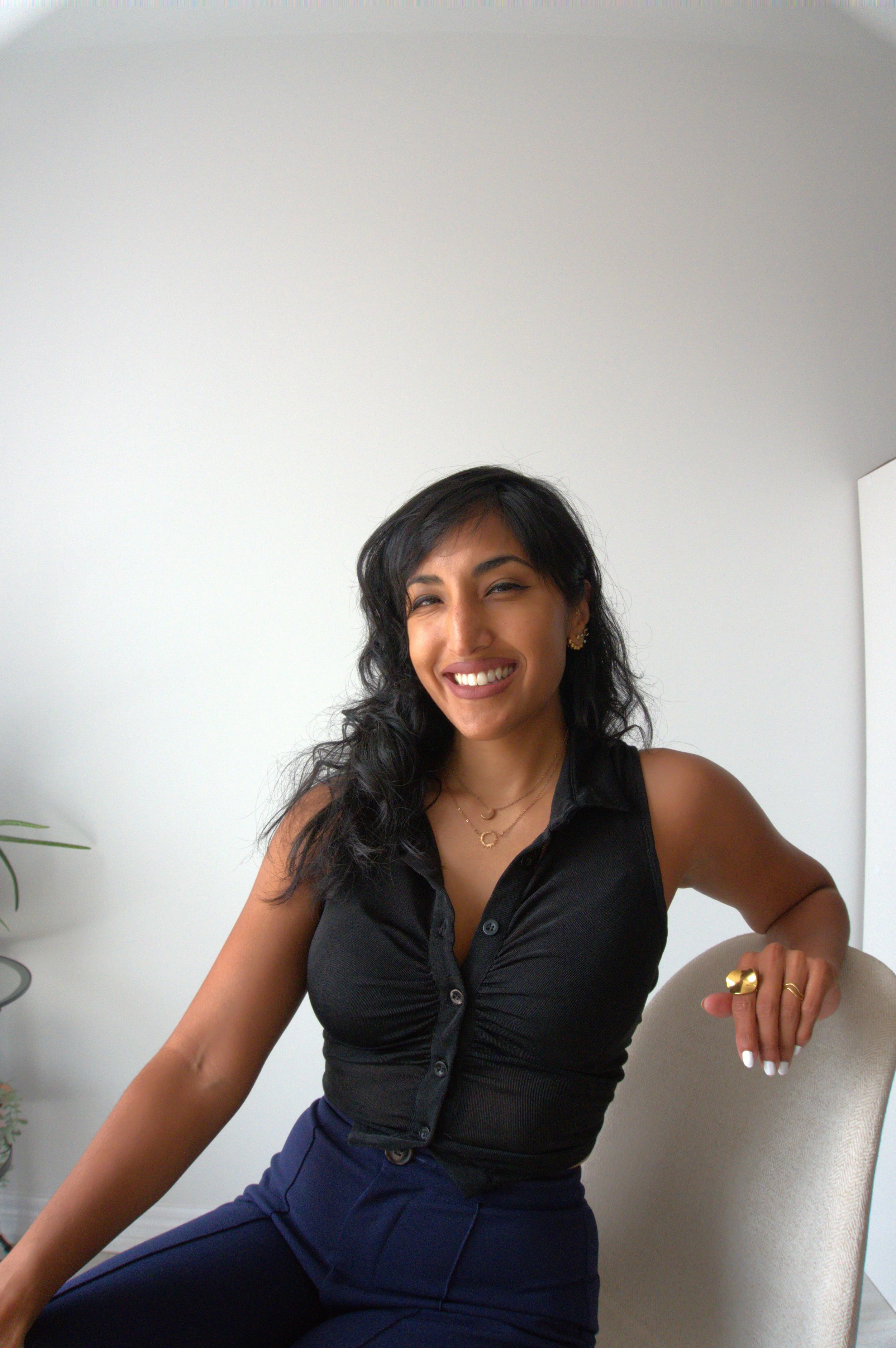 A woman with dark, wavy hair smiling, sitting on a beige chair, wearing a sleeveless black top with buttons, blue pants, and gold jewelry, in a bright room with white walls and a small plant on a side table.