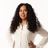 A woman with curly hair wearing a white blouse, standing with her hand on her hip, facing forward.