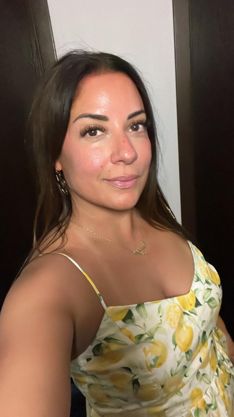 A woman with medium brown hair styled straight, wearing a yellow and white lemon-patterned dress with spaghetti straps, gold earrings, a delicate gold necklace, and light makeup, standing in front of a dark door and a white wall.