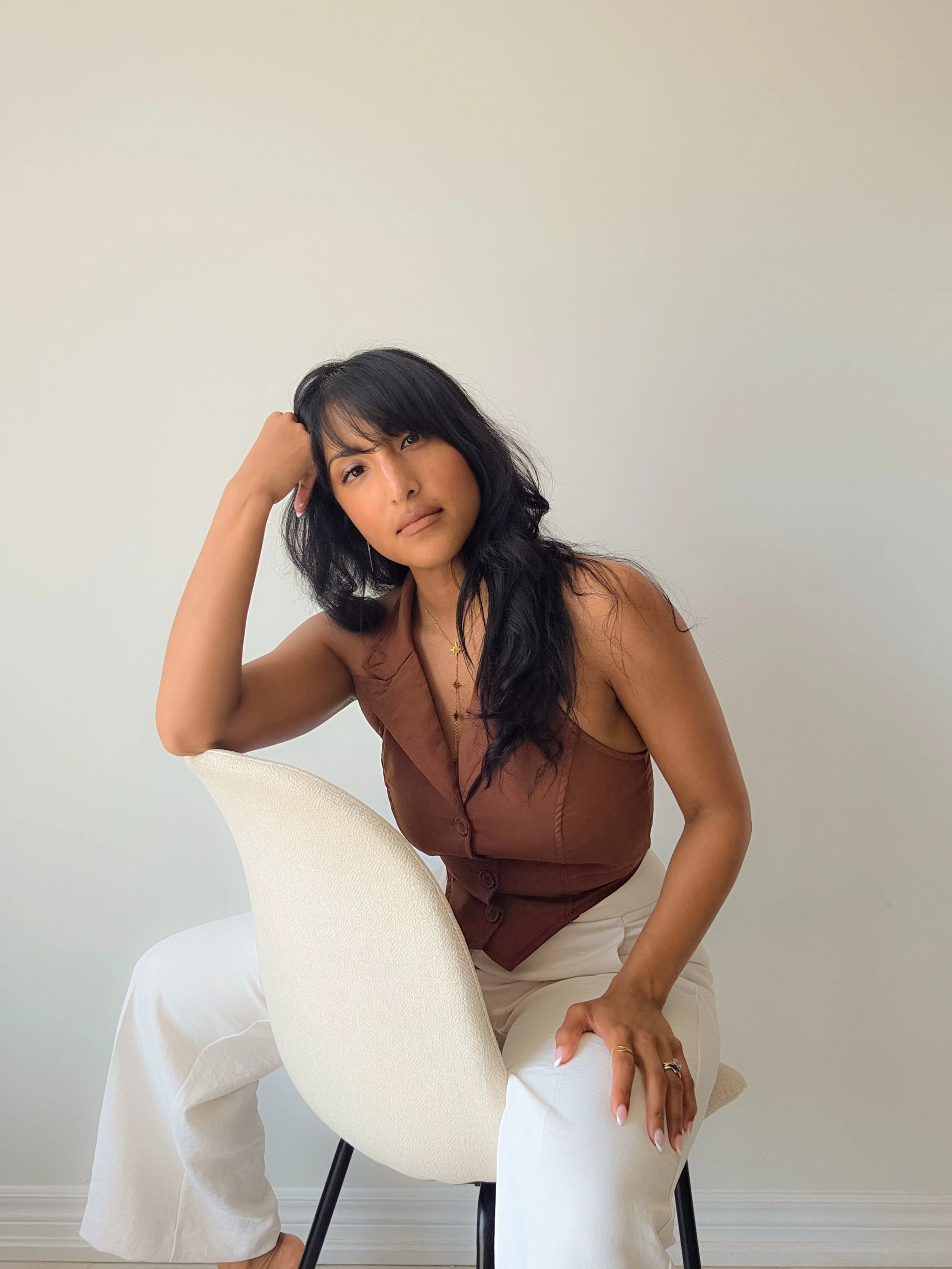 Woman with long dark hair, wearing a brown sleeveless top and white pants, sitting on a white chair against a plain light background, resting her head on her hand with an expression of calm and contemplation.