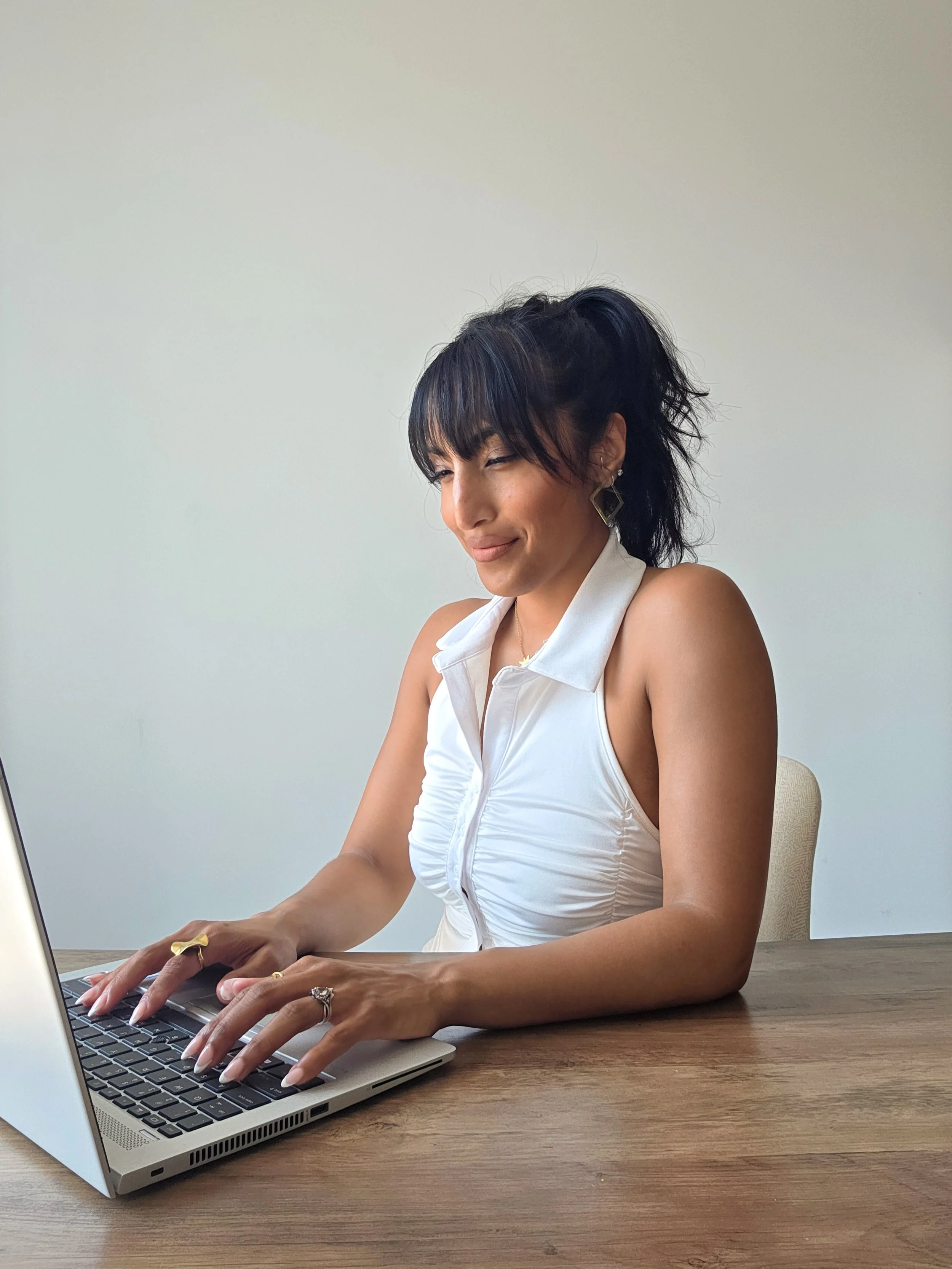 A woman with dark, curly hair styled in a high ponytail, wearing a white sleeveless top, large geometric earrings, and several rings, sitting at a wooden table typing on a laptop with a neutral white wall in the background.