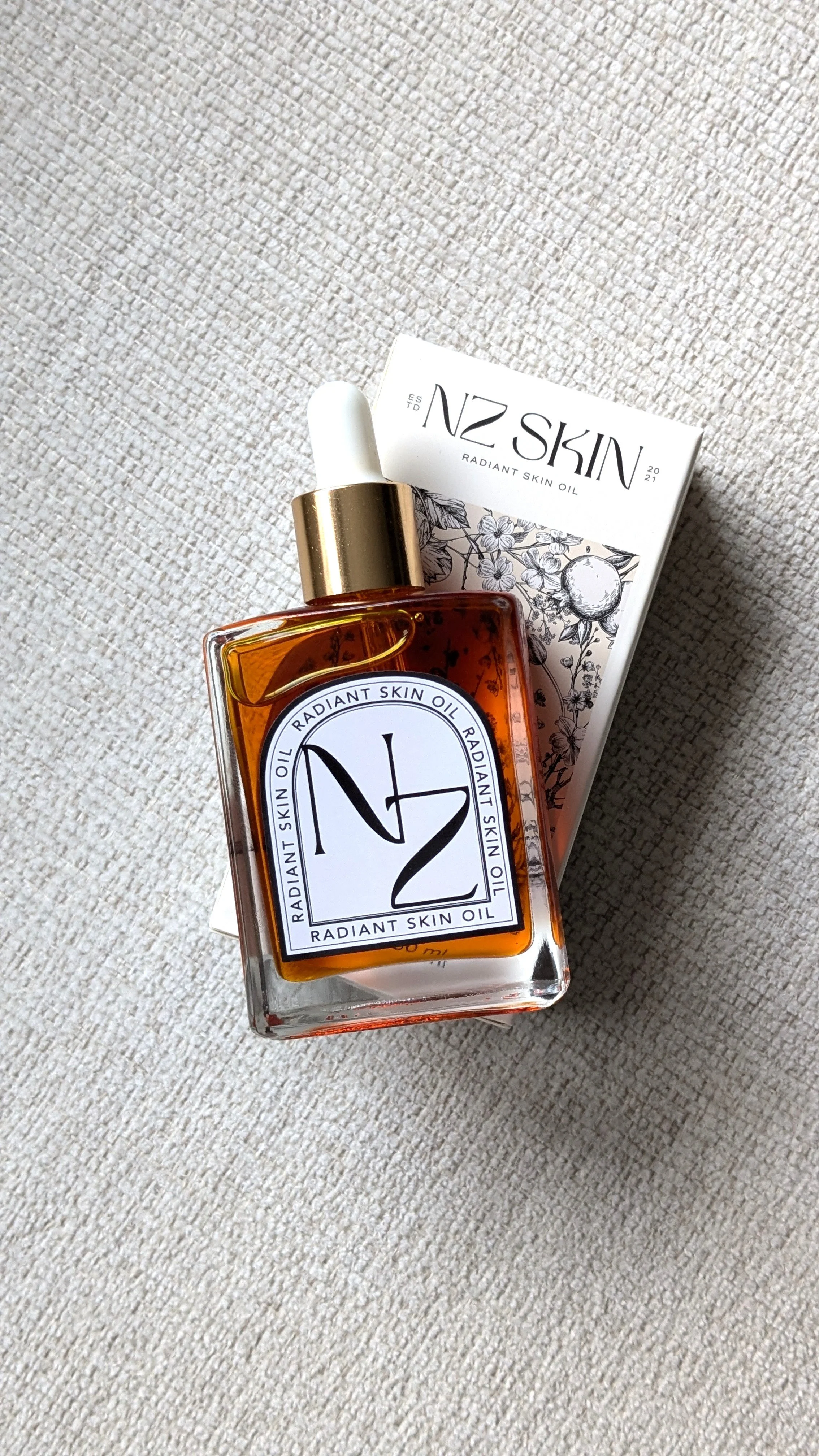 A bottle of NZ Skin Radiant Skin Oil with a white dropper cap, placed on top of its packaging box, on a textured beige surface.