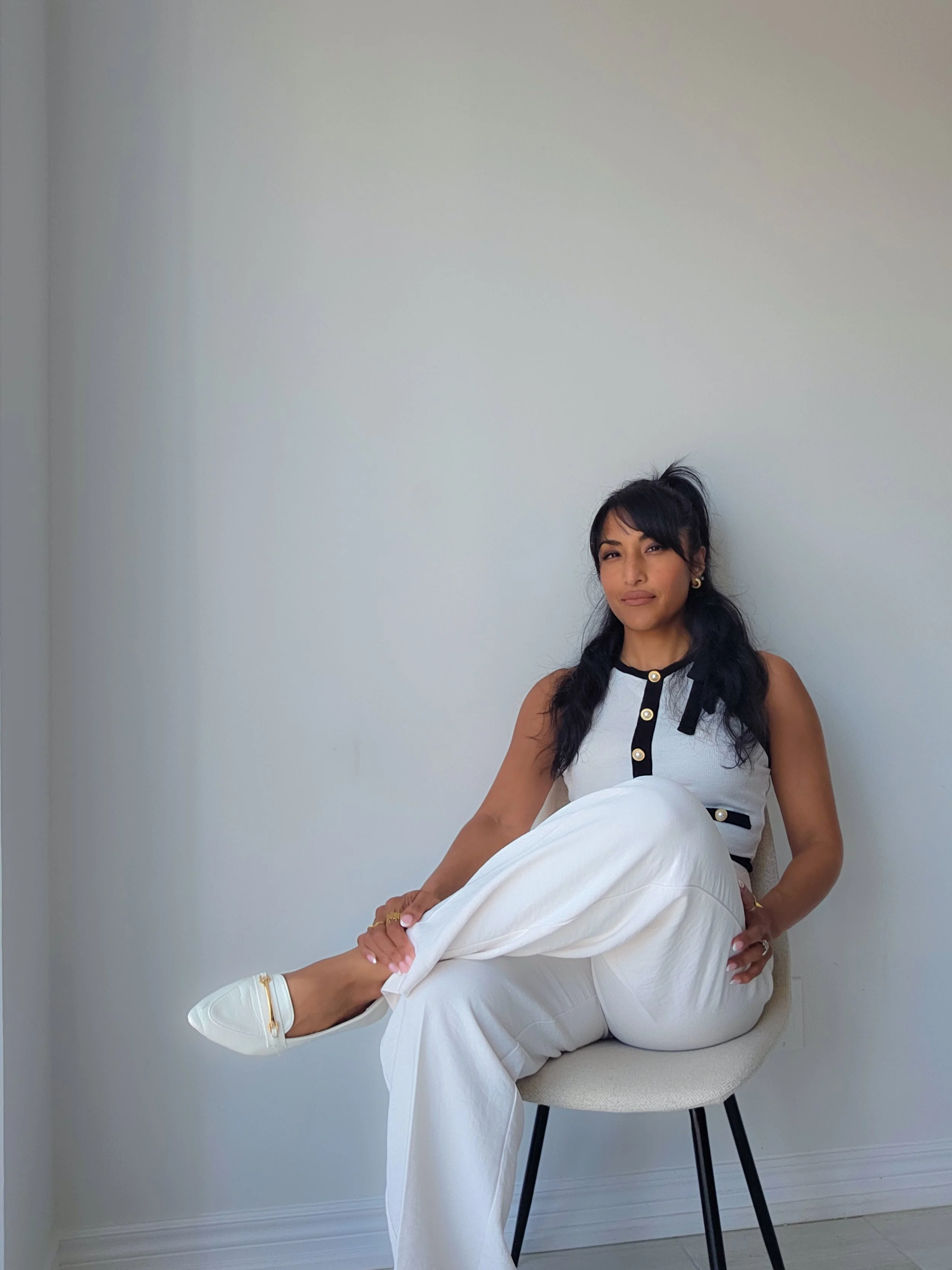 Two women sitting on a beige chair in front of a plain white wall, one with long black hair and the other with short black hair and bangs, dressed in casual white and black clothing.