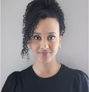 A woman with curly hair pulled to one side, wearing a black top, facing the camera with a neutral background.