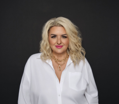 A woman with blonde curly hair wearing a white button-up shirt and gold jewelry against a black background.