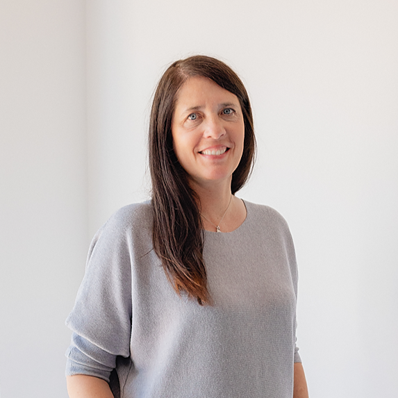 Woman with long dark hair wearing a light gray sweater and standing in front of a white wall.