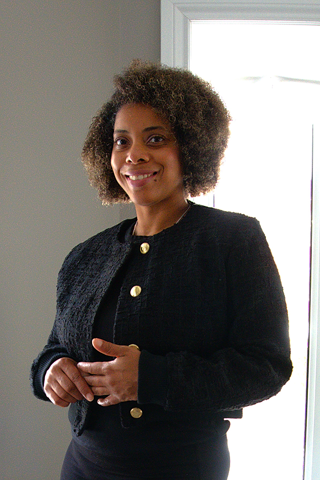A woman with curly hair and a black outfit stands indoors near a window, smiling.