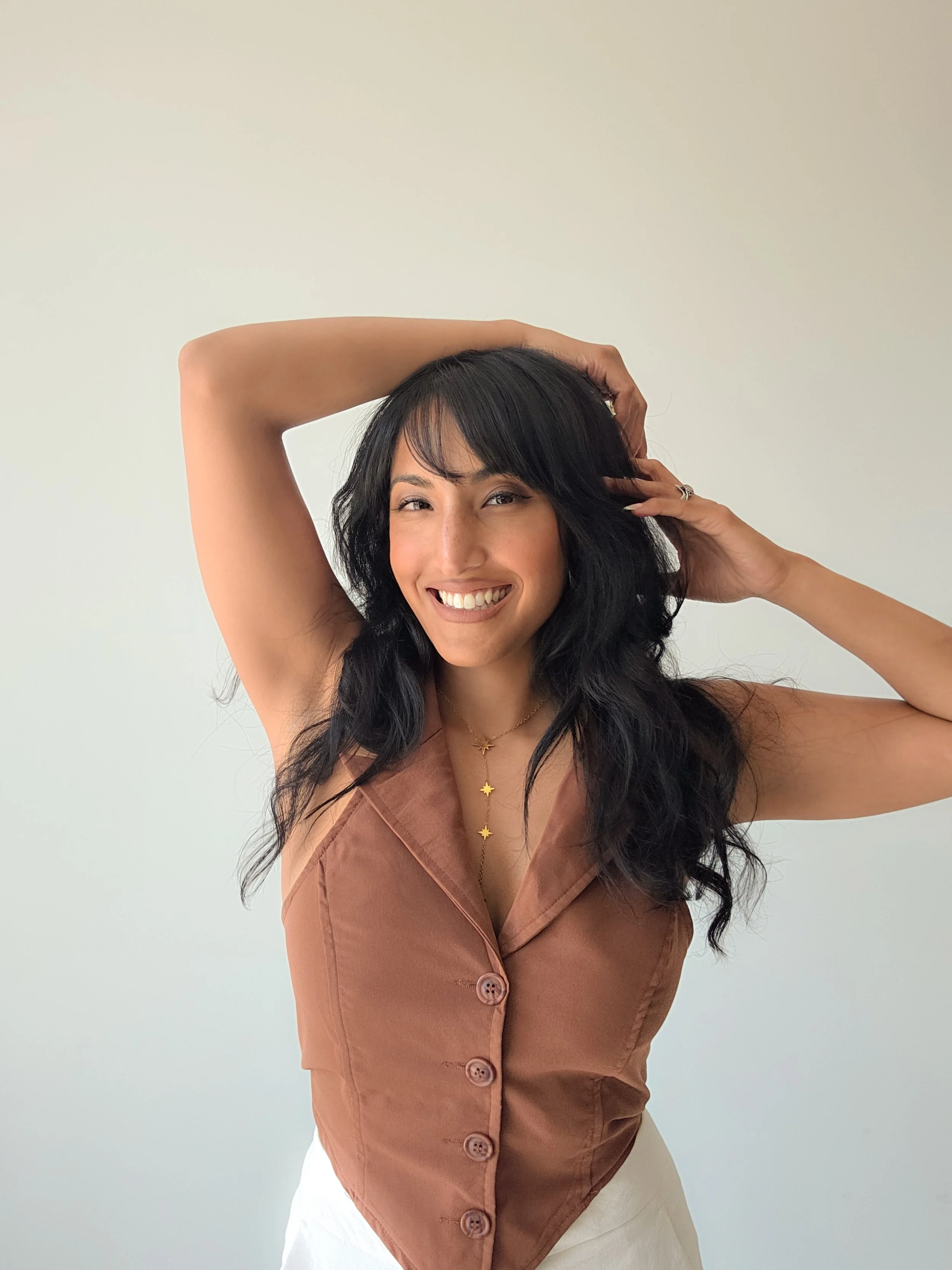 A woman with long black hair and light skin smiling, wearing a sleeveless brown button-up top and white pants, posing with hands behind her head in front of a plain light-colored wall.