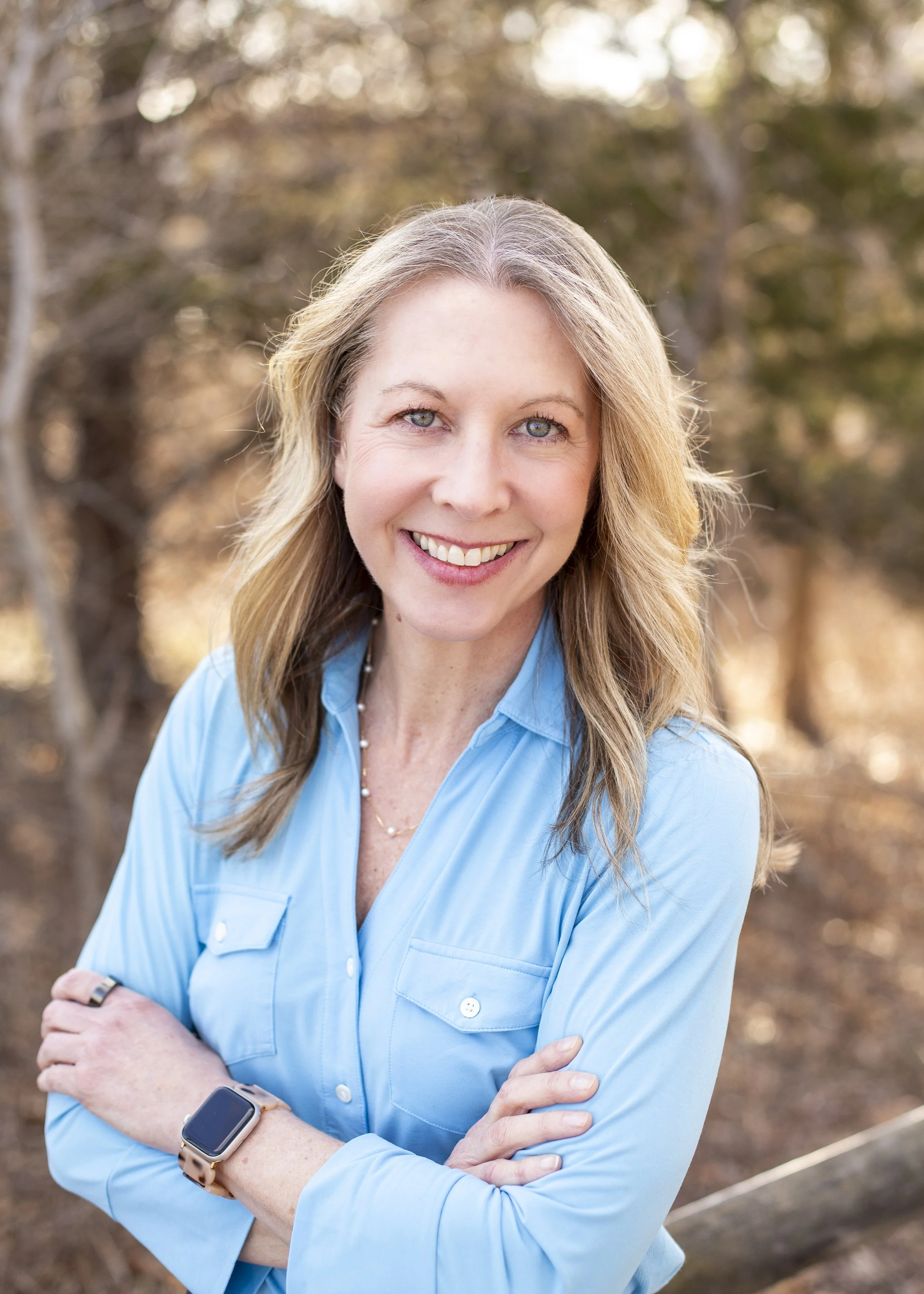 A woman with blonde hair smiling, wearing a light blue button-up shirt, standing outdoors with blurred trees in the background.