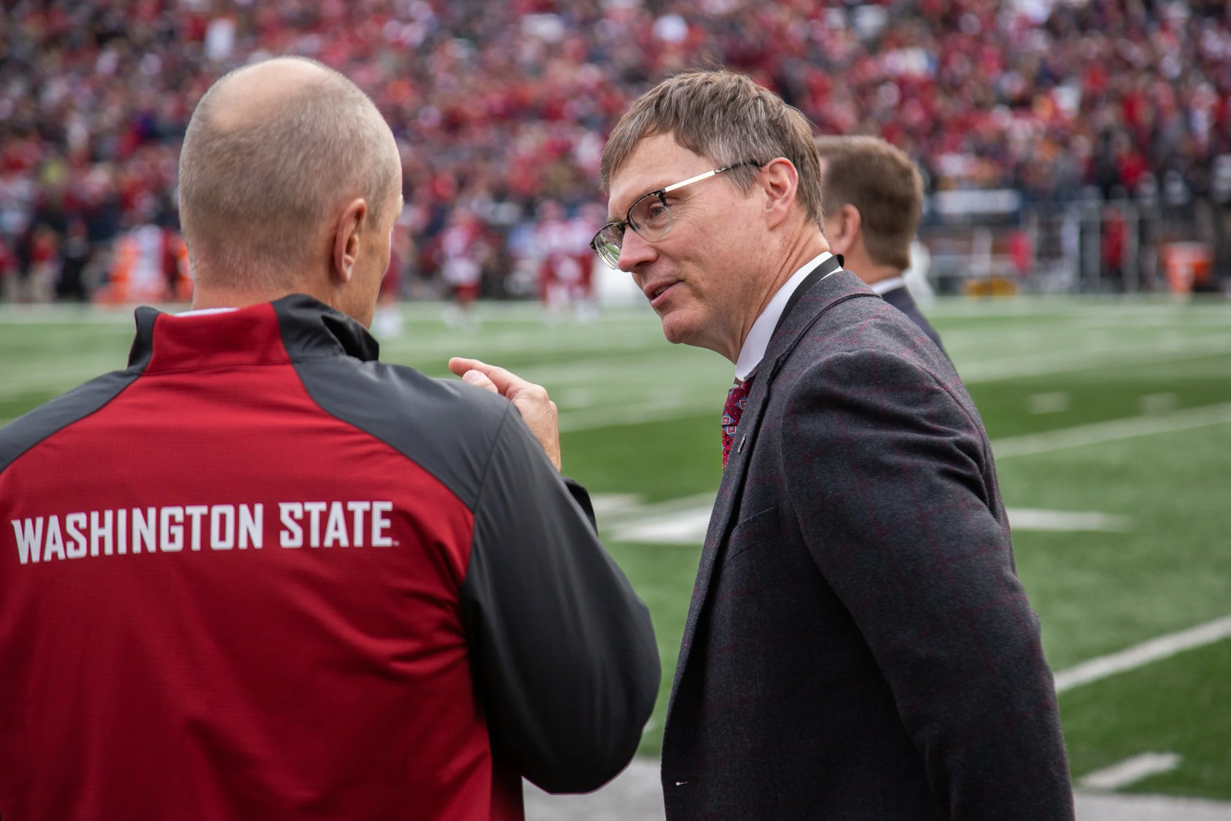 Two men having a conversation on a football field, one wearing a red Washington State jacket and the other in a gray suit with glasses, with a stadium full of spectators in the background.