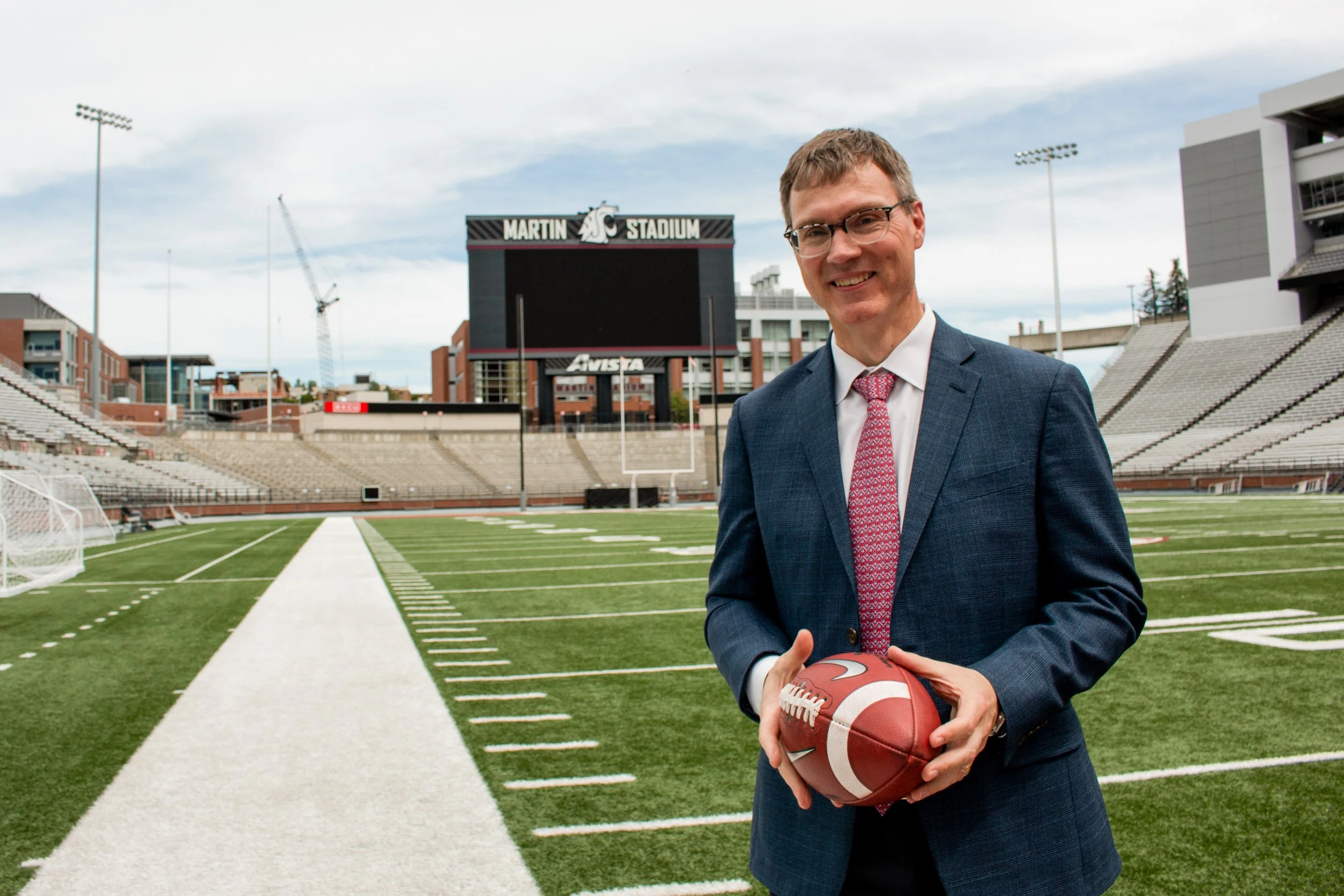 A man in a suit holding an American football in a stadium with empty seats, a large scoreboard reading 'Martin Stadium,' and overcast skies overhead.