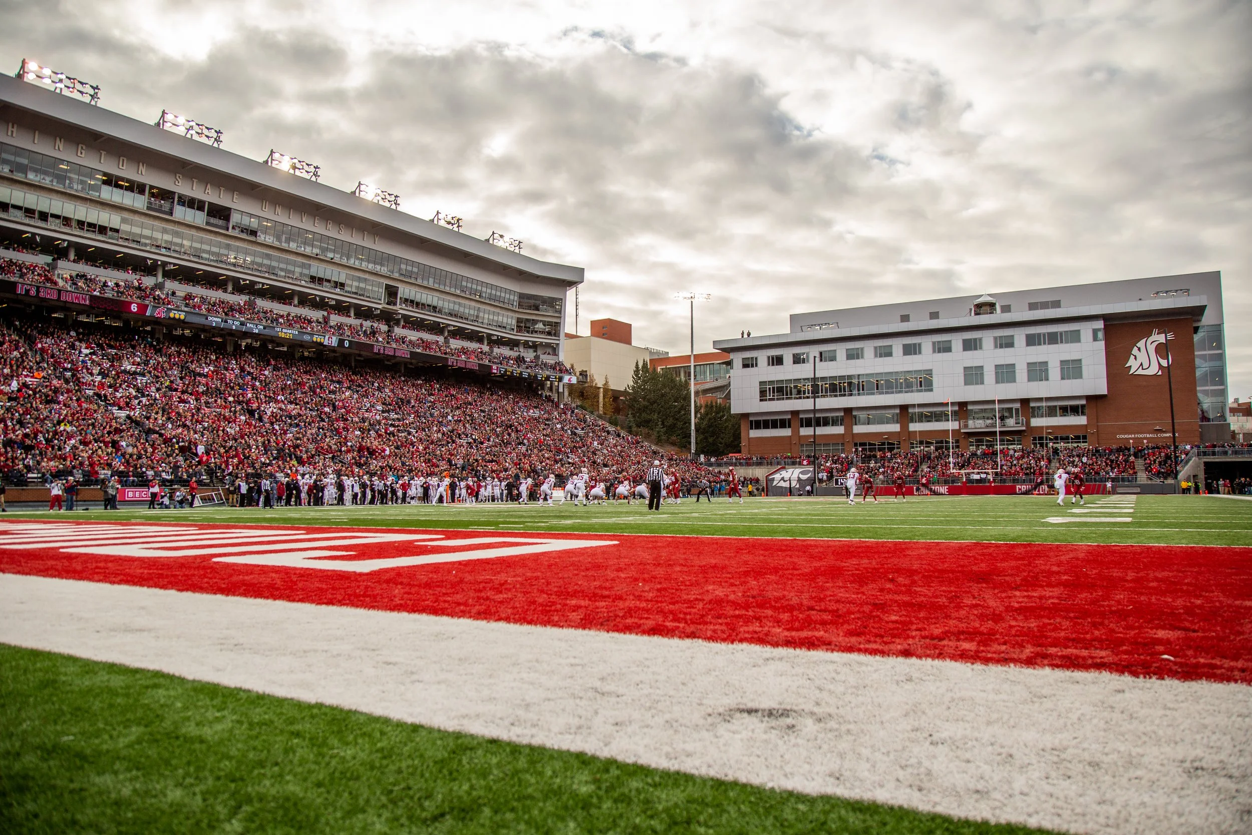 Wide view of a college football game at Washington State University, with spectators filling the stadium, players on the field, and a cloudy sky overhead.