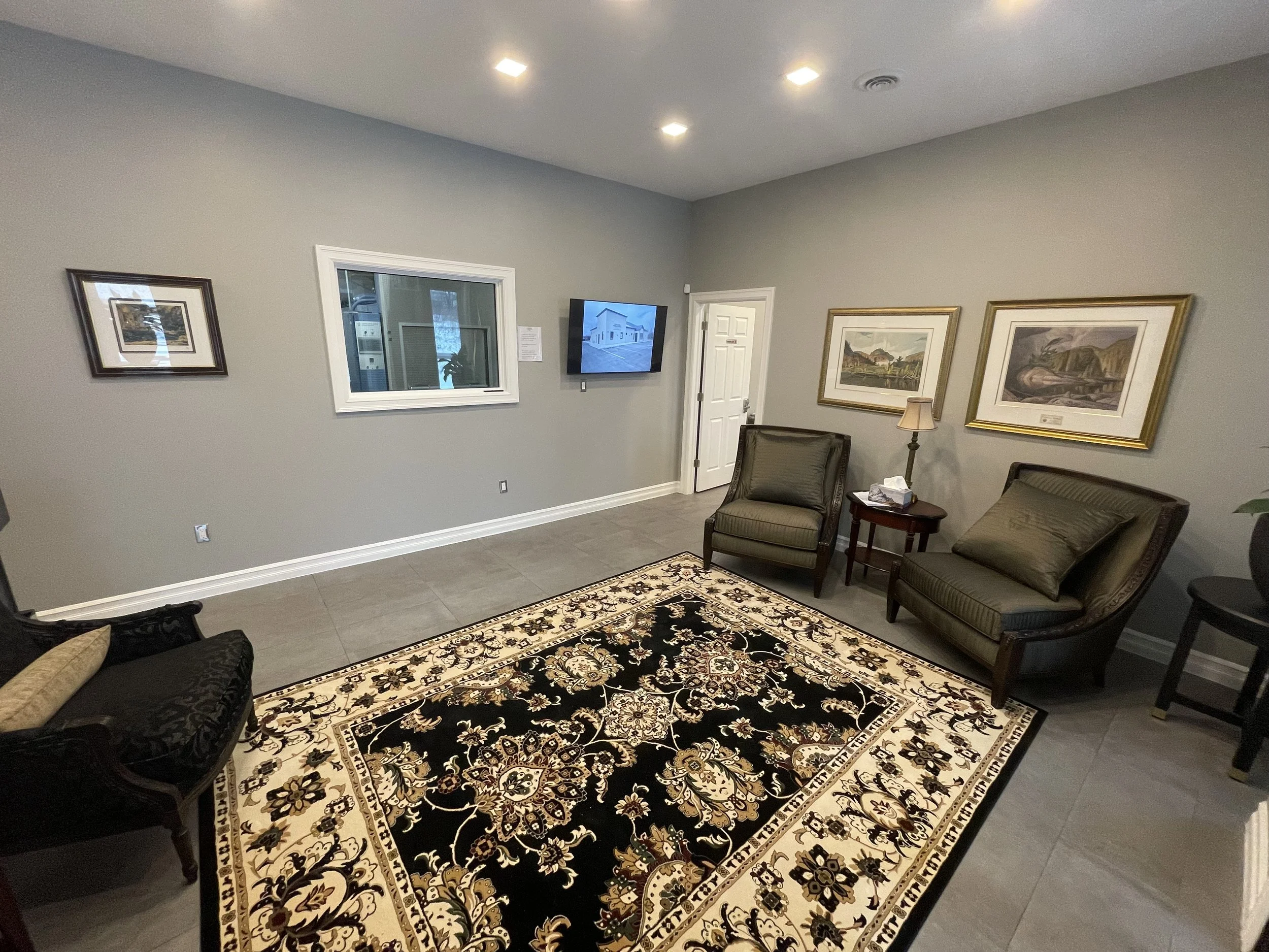 A waiting room with gray walls, decorated with framed landscape artwork, and a patterned black, white, and beige rug. There are two green armchairs, a small wooden side table with tissues, and a wall-mounted TV showing an image of a building. A window and a white door are visible, with ceiling lights illuminating the room.