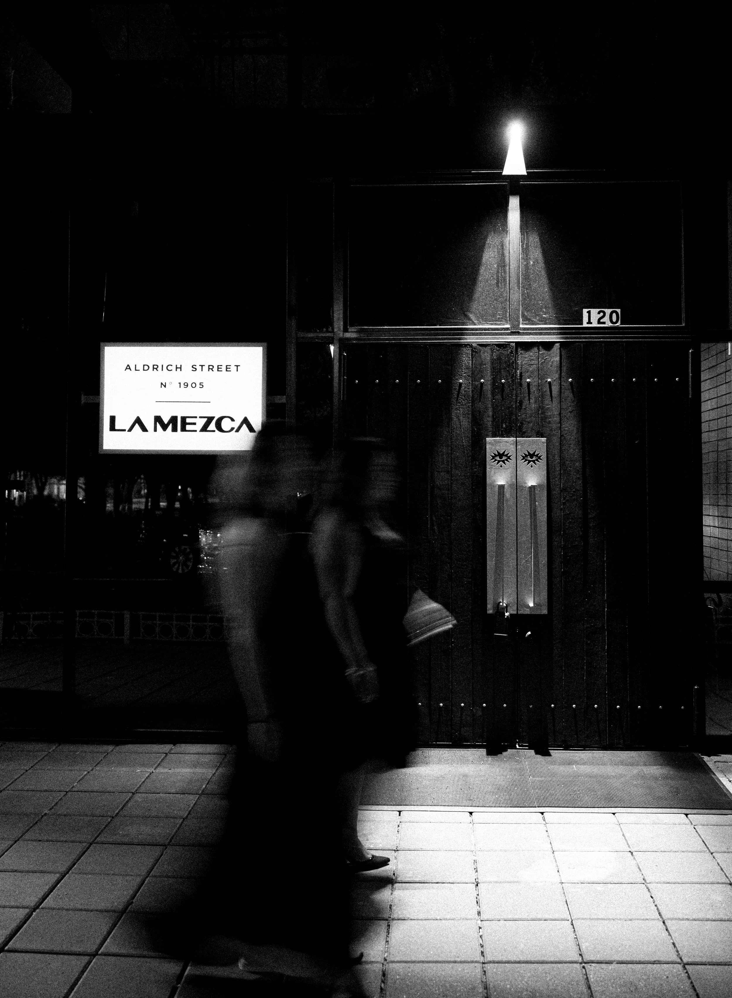 Exterior of La Mezca bar at night showing the illuminated entrance sign and two decorative stone door panels with eye motifs