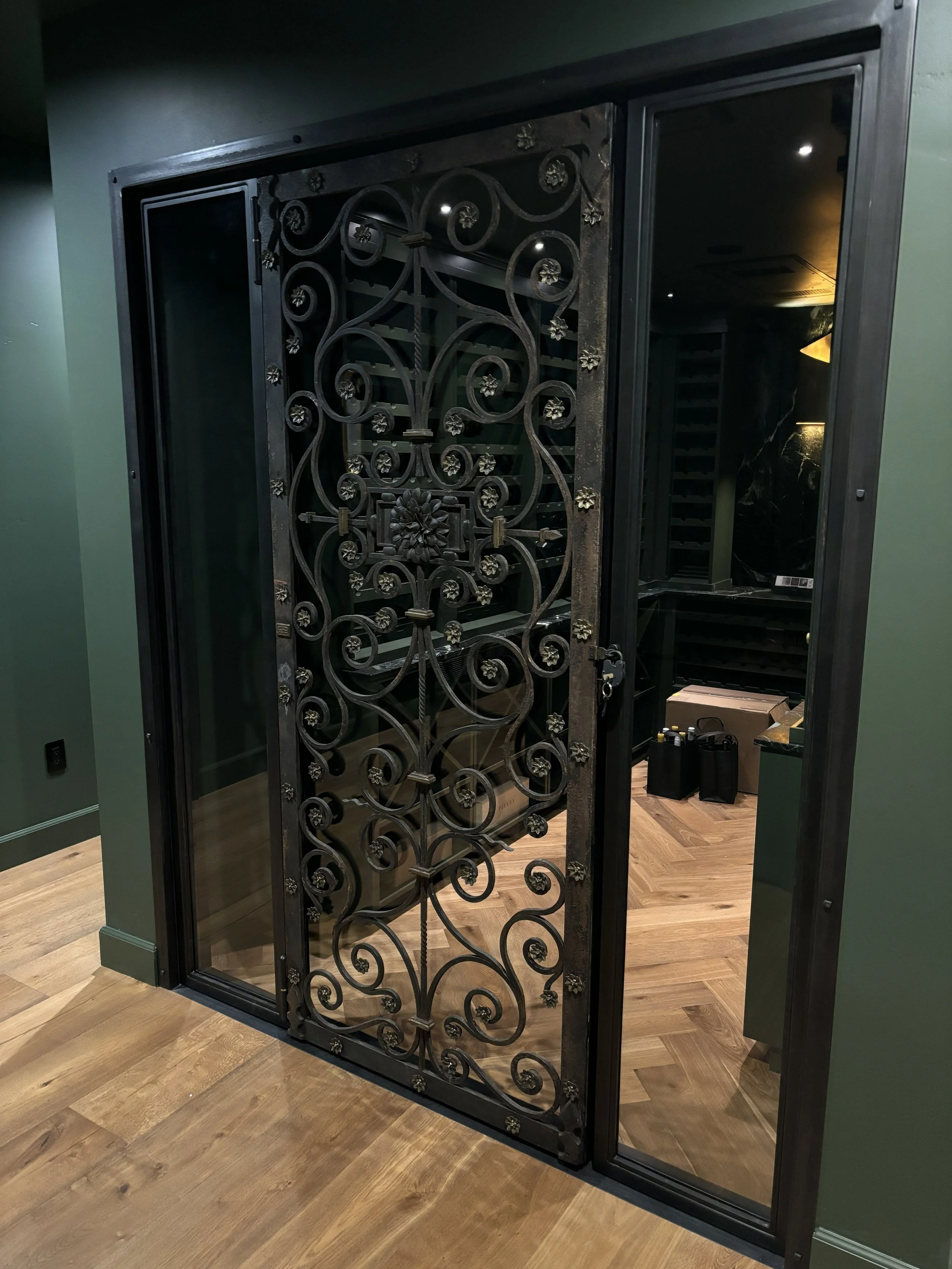 Decorative black wrought iron and glass door with intricate scrollwork and floral details leading into a wine cellar or storage room.
