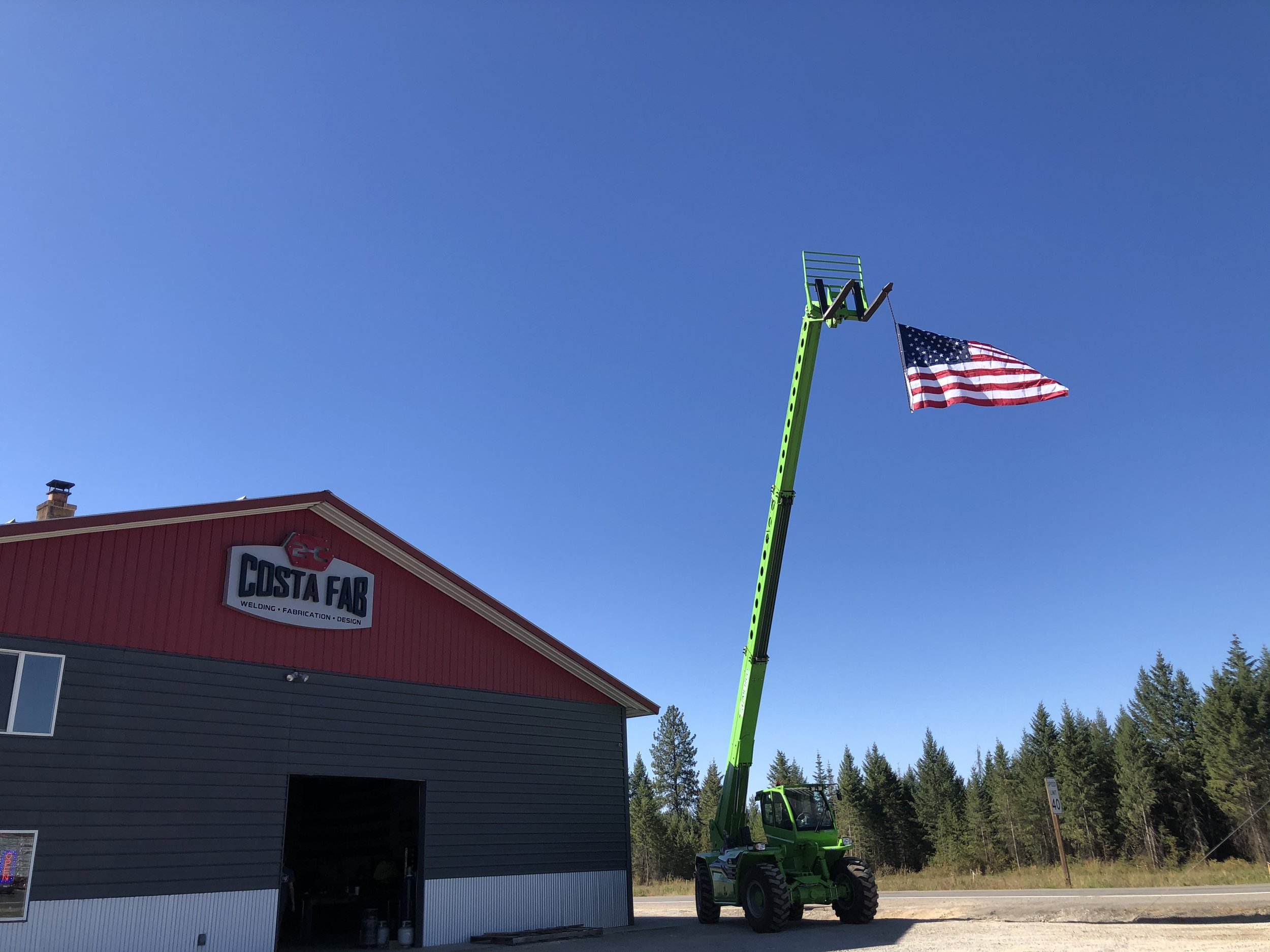 A green lift is extending to hold an American flag near a red and gray building with a sign reading 'Costa Far'. The sky is clear with no clouds, and a line of trees is visible in the background.