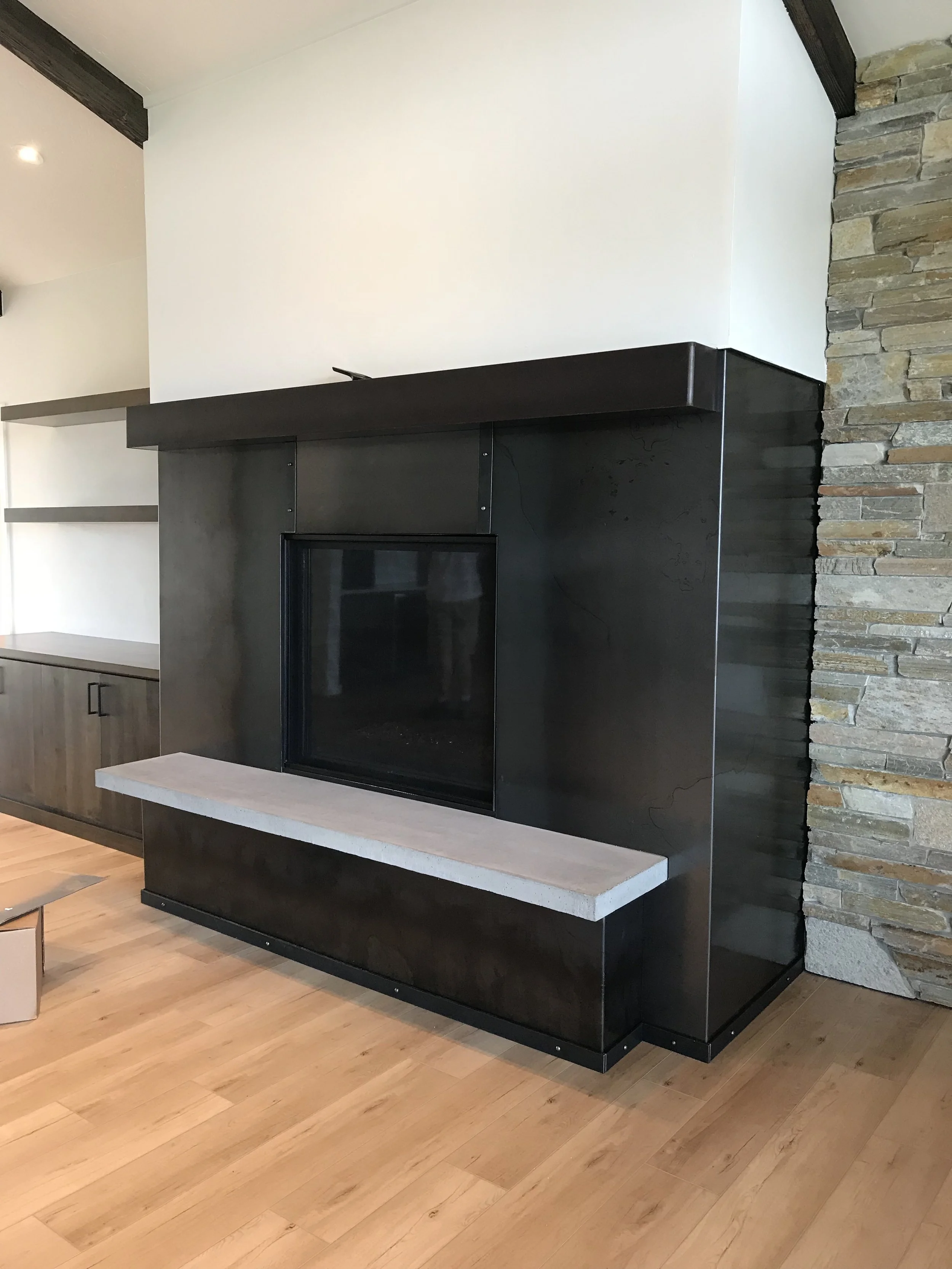 Modern living room fireplace with black metal surround, stone wall on the right, and light wood flooring in the foreground.