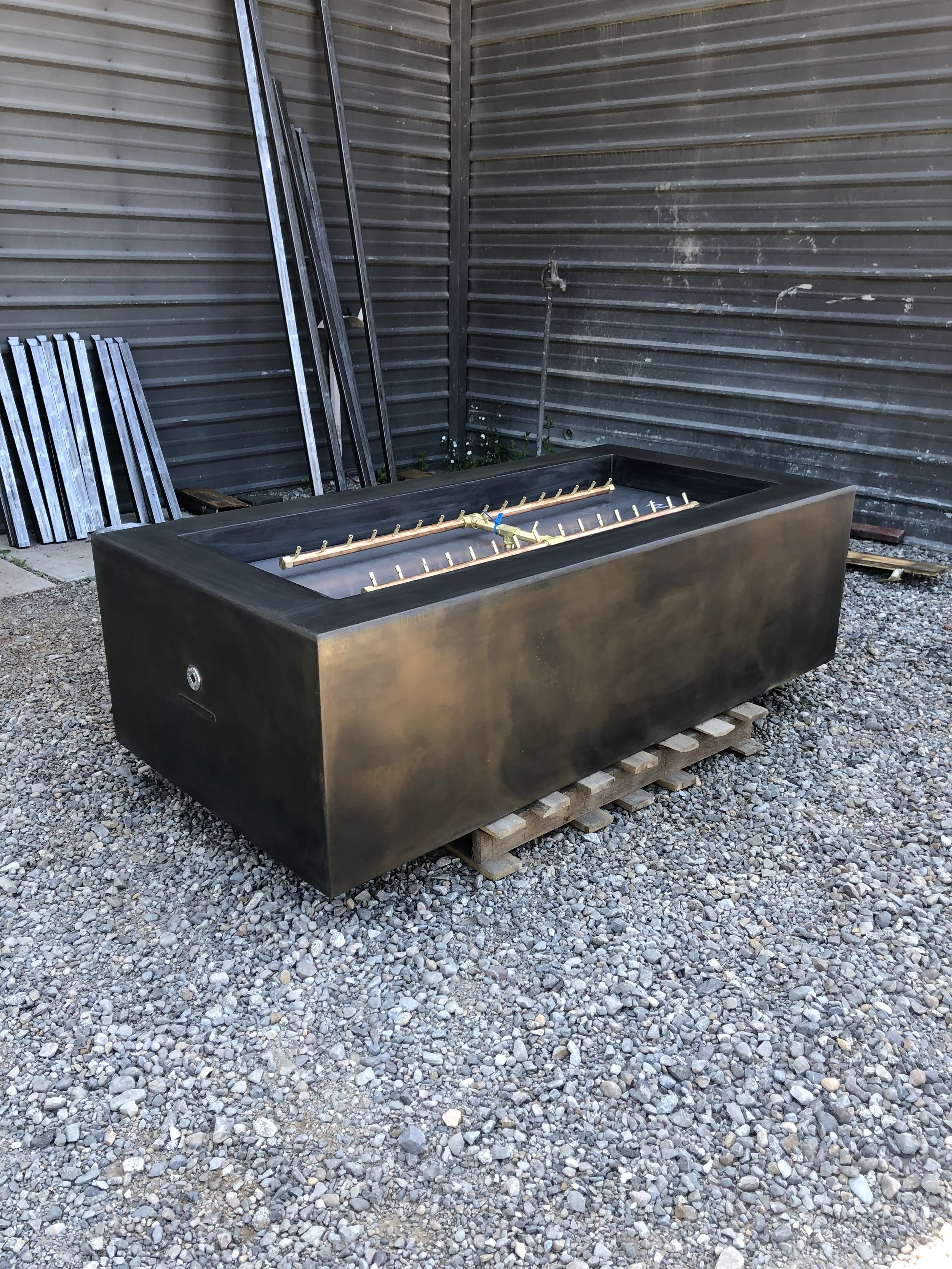An outdoor hot tub with a black exterior and multiple jets, empty and covered with a black cover, situated on a wooden pallet on gravel with metal fencing in the background.