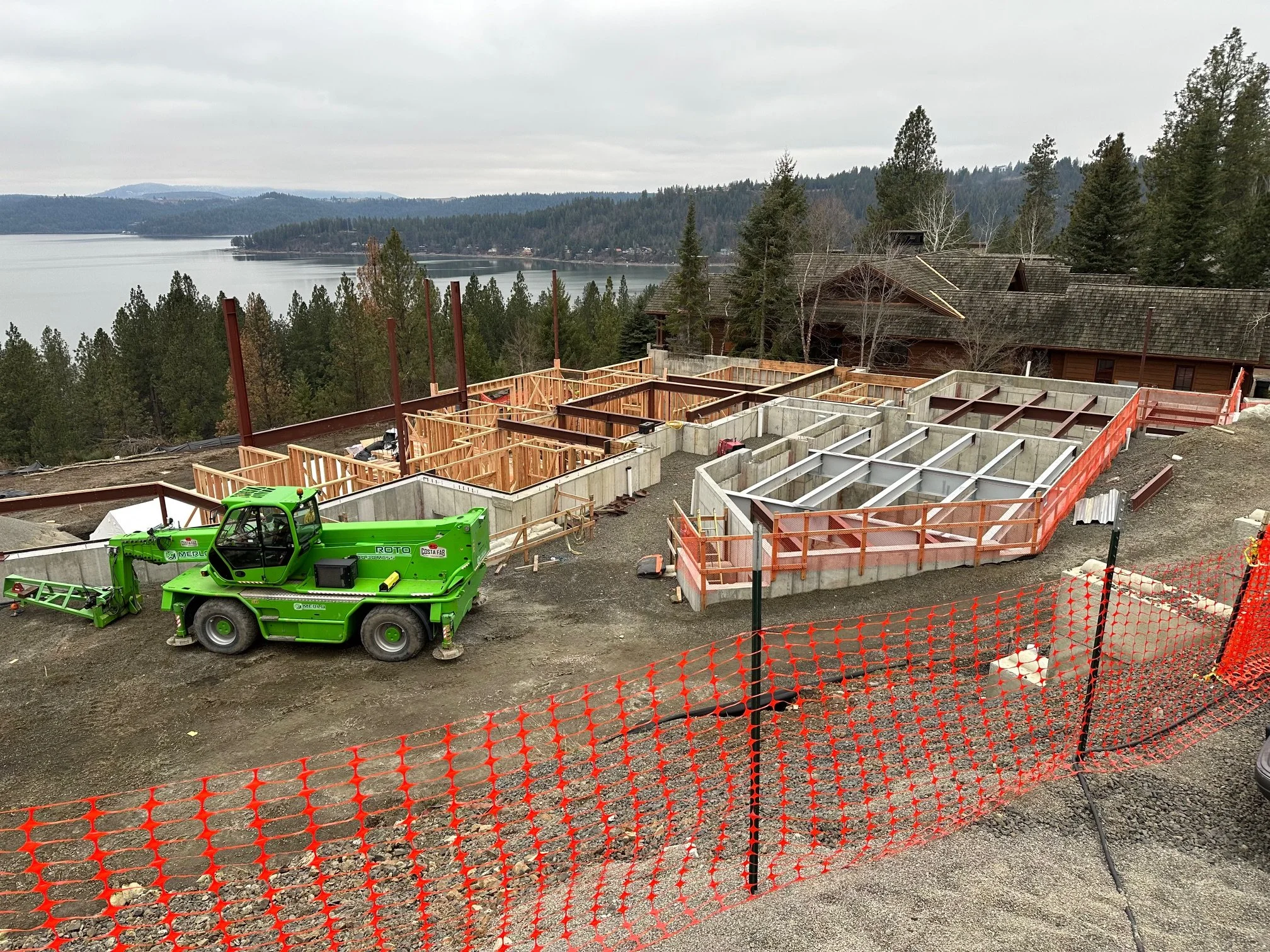Construction site on a hillside overlooking a lake, with a bright green construction vehicle and partially built foundation walls with framing, orange safety barriers, and trees in the background.