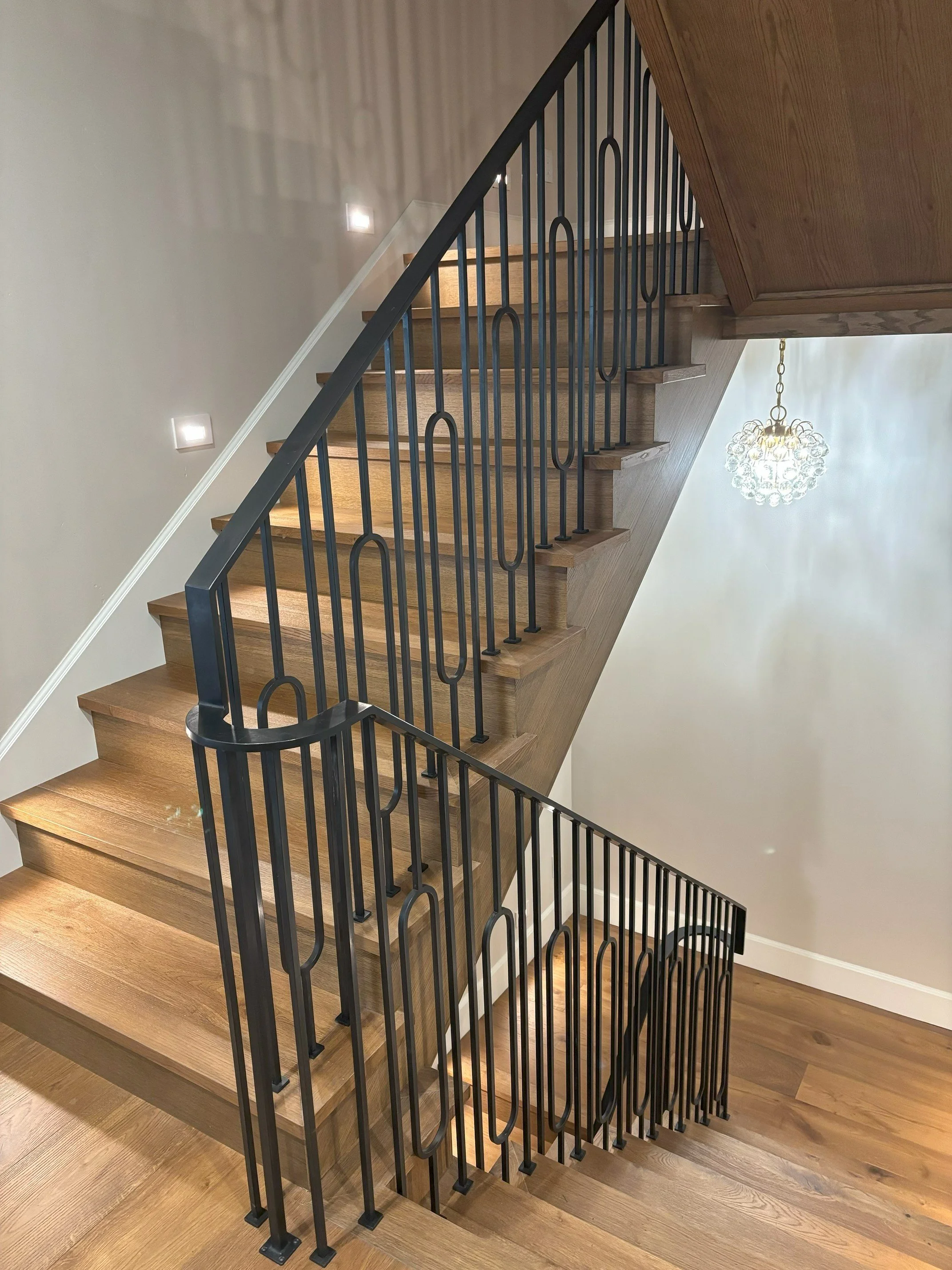 Interior view of a modern wooden staircase with black metal railings, ascending from a wooden floor, illuminated by small wall lights and a chandelier hanging from the ceiling.