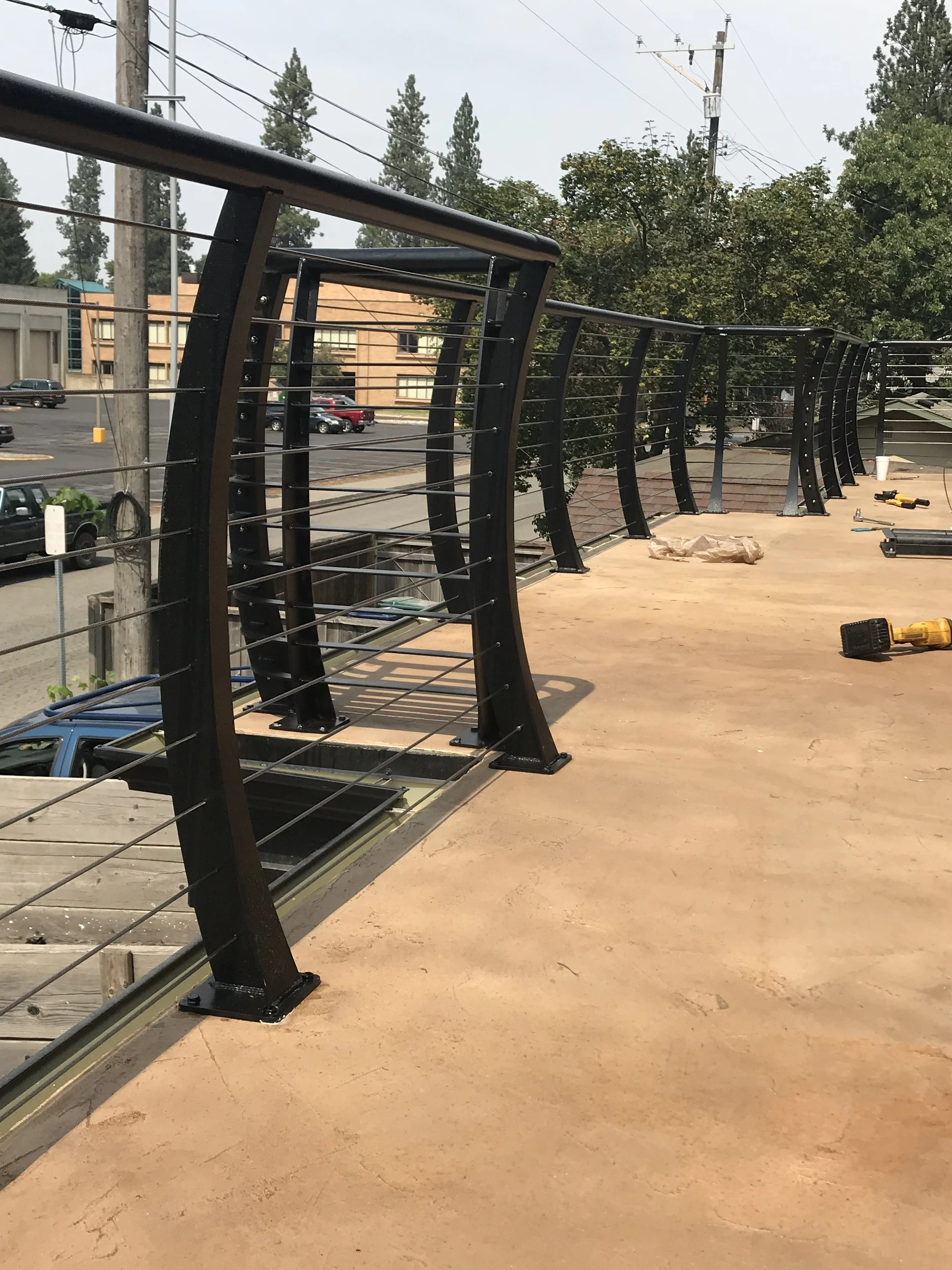 View of a balcony or terrace under construction with black metal railing, wooden floor, and tools scattered around. Trees, power lines, and parked cars are visible in the background.