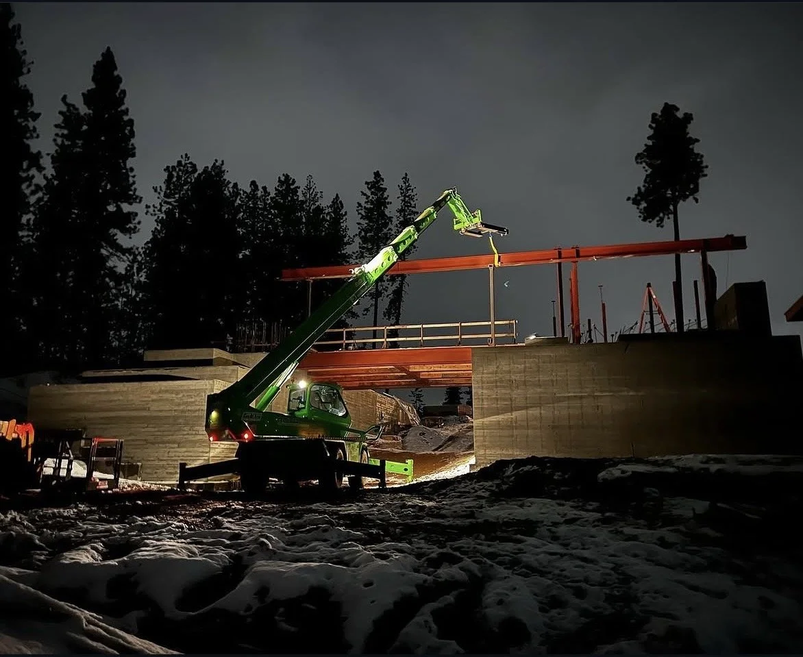 Construction site at night with a green crane lifting a beam, snow on the ground, and trees in the background.