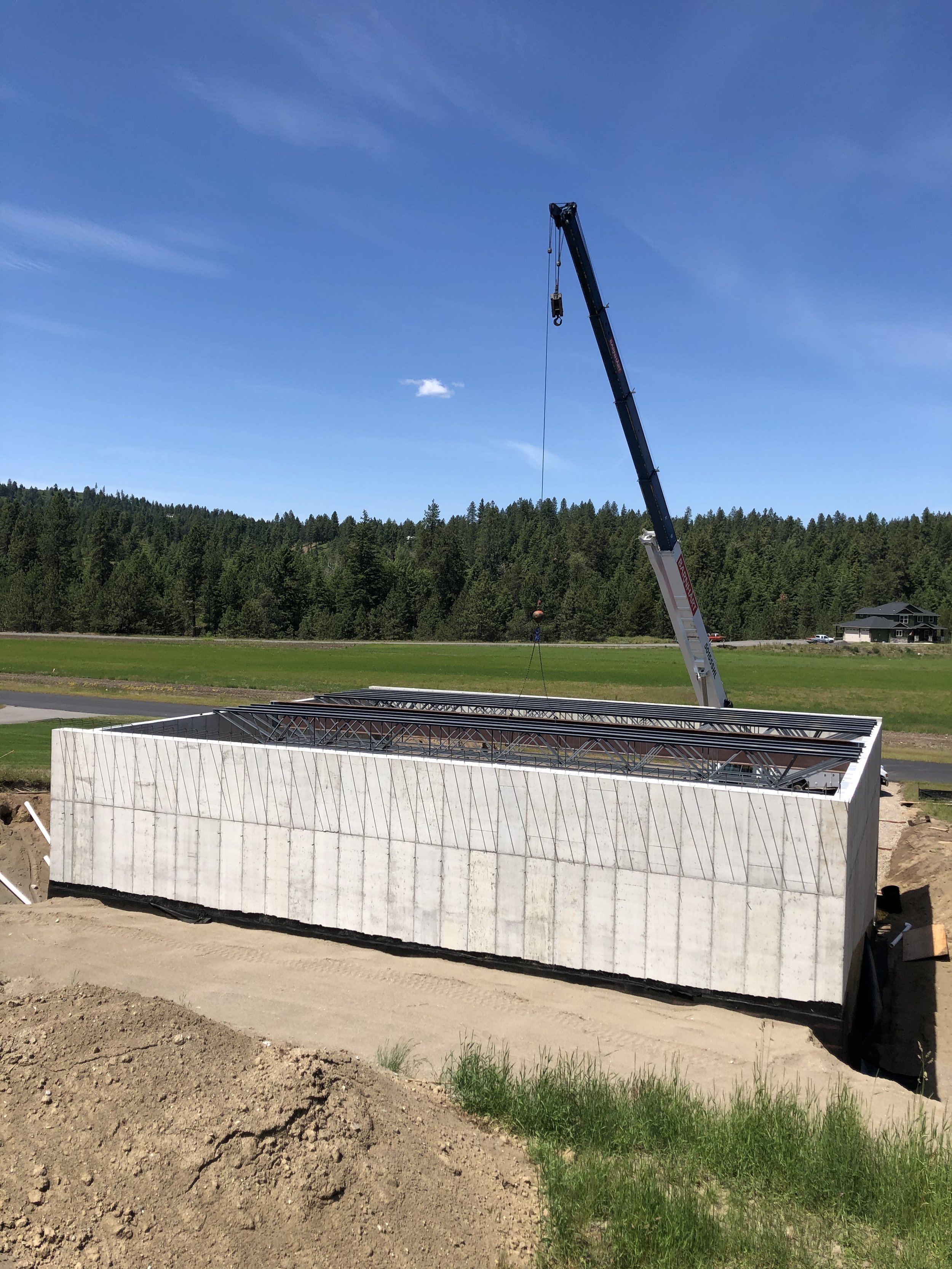 Construction site with a large rectangular building foundation, a crane lifting steel beams, and a background of green trees and a blue sky.