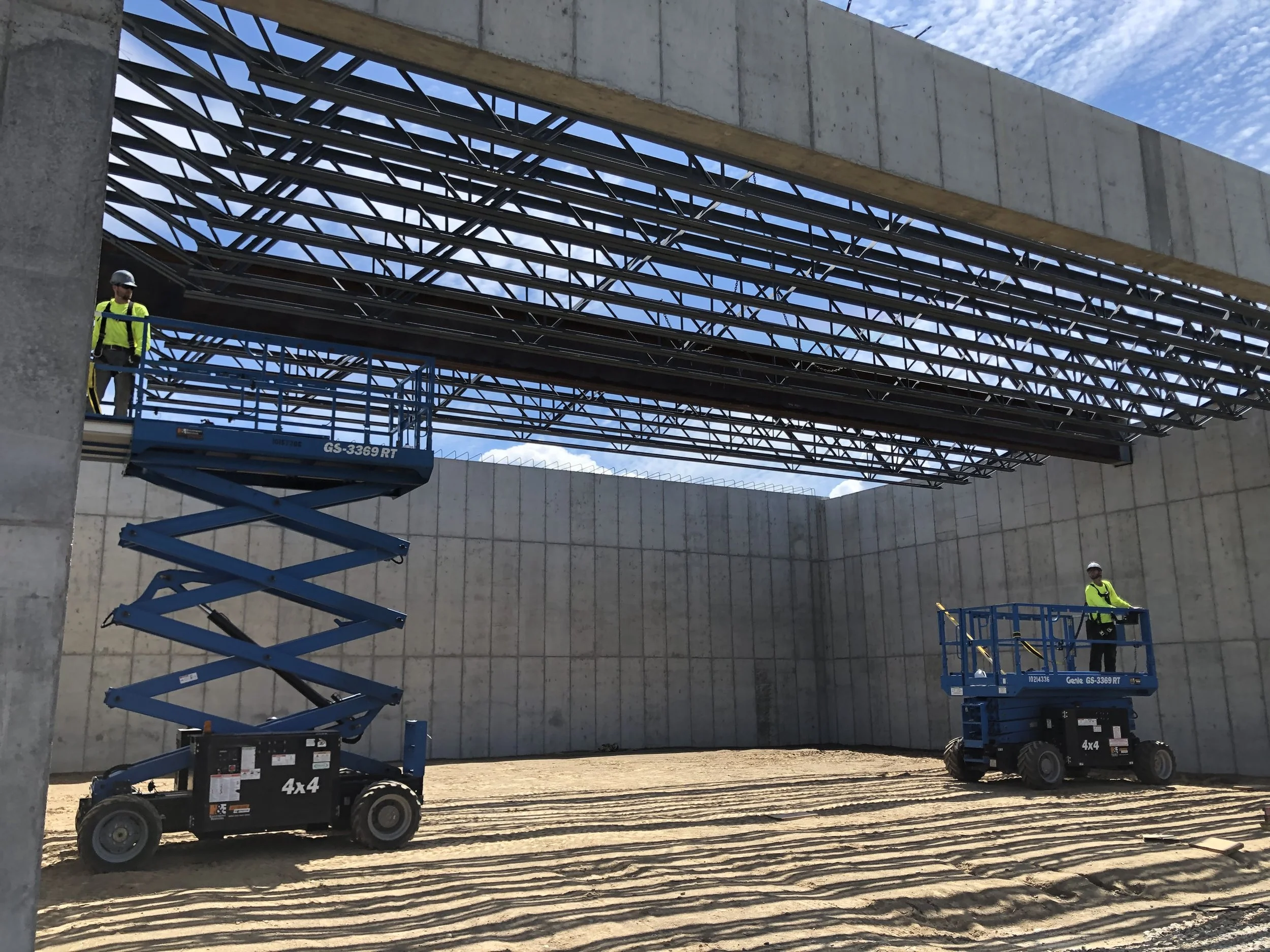 Two construction workers on blue scissor lifts working on a large metal structure above an unfinished concrete space with dirt ground.