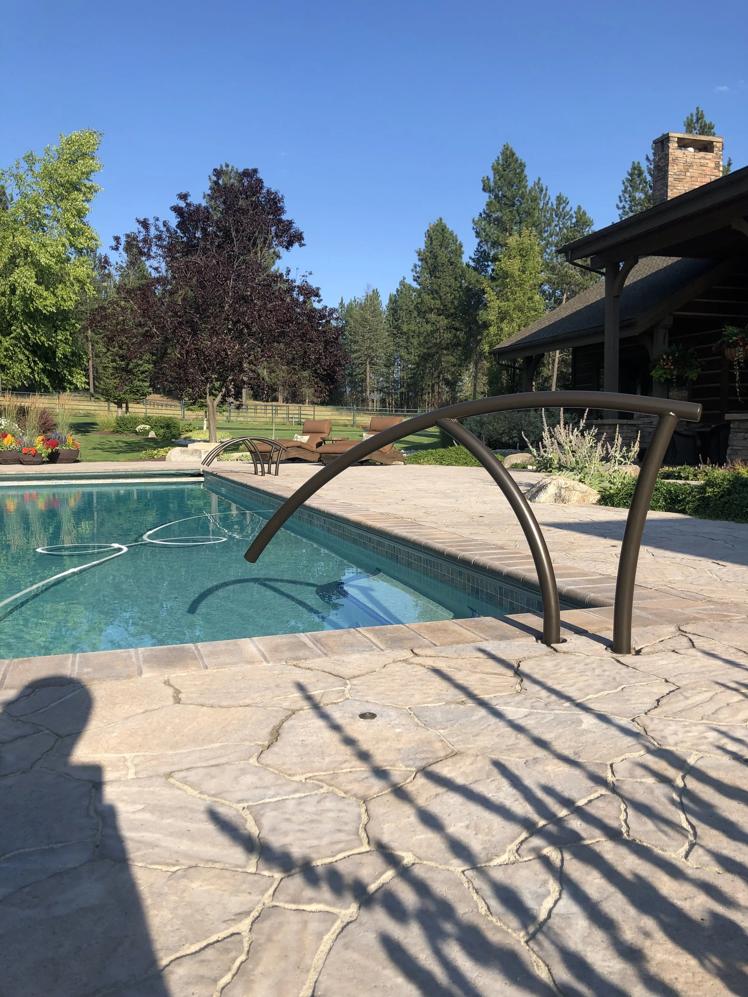 Photo of a backyard with a swimming pool, stone patio, and trees under a clear blue sky.