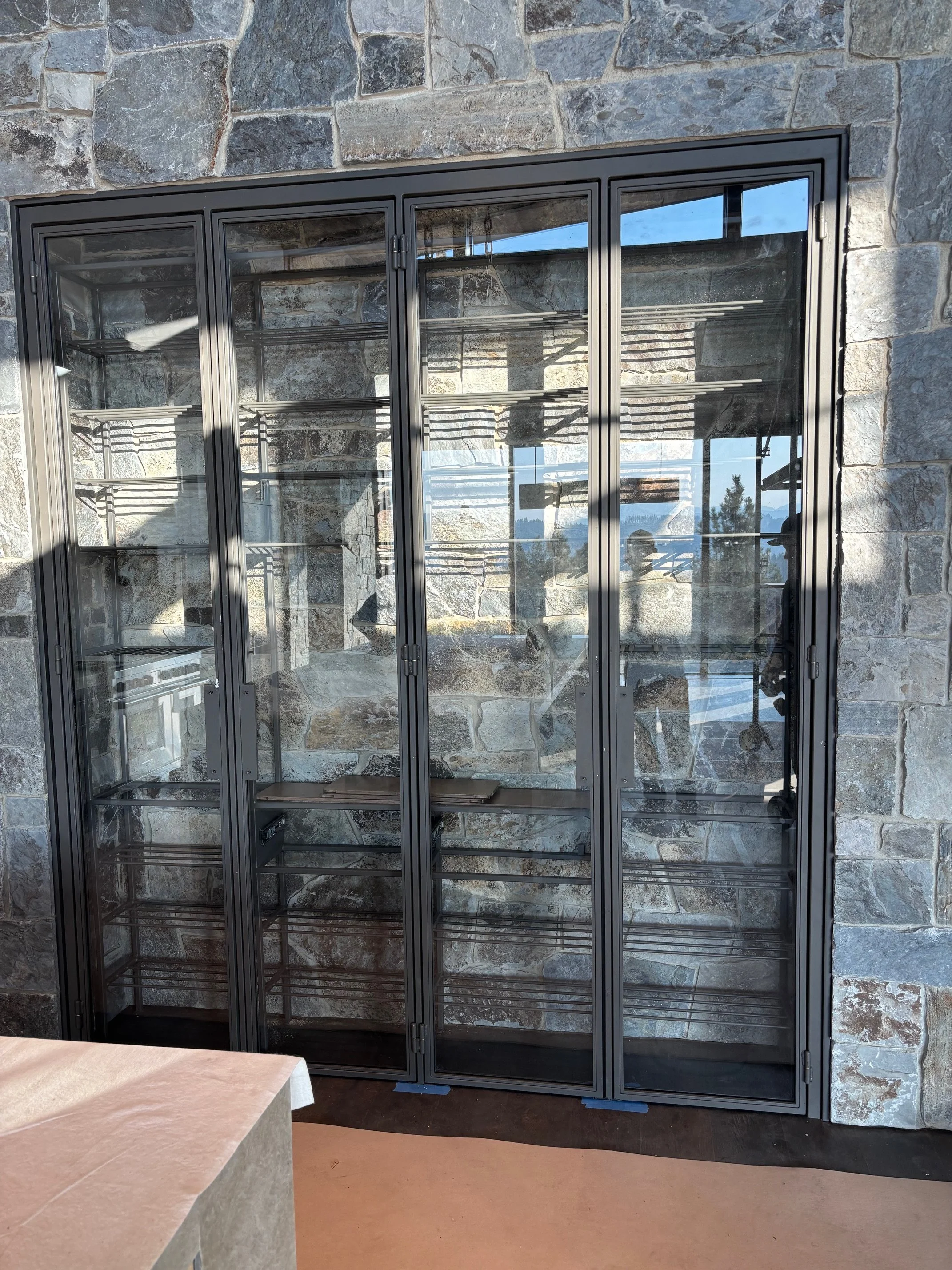 Empty glass display cabinet with black metal frame in front of a stone wall, with multiple shelves inside and reflection of outdoor scenery in the glass doors.