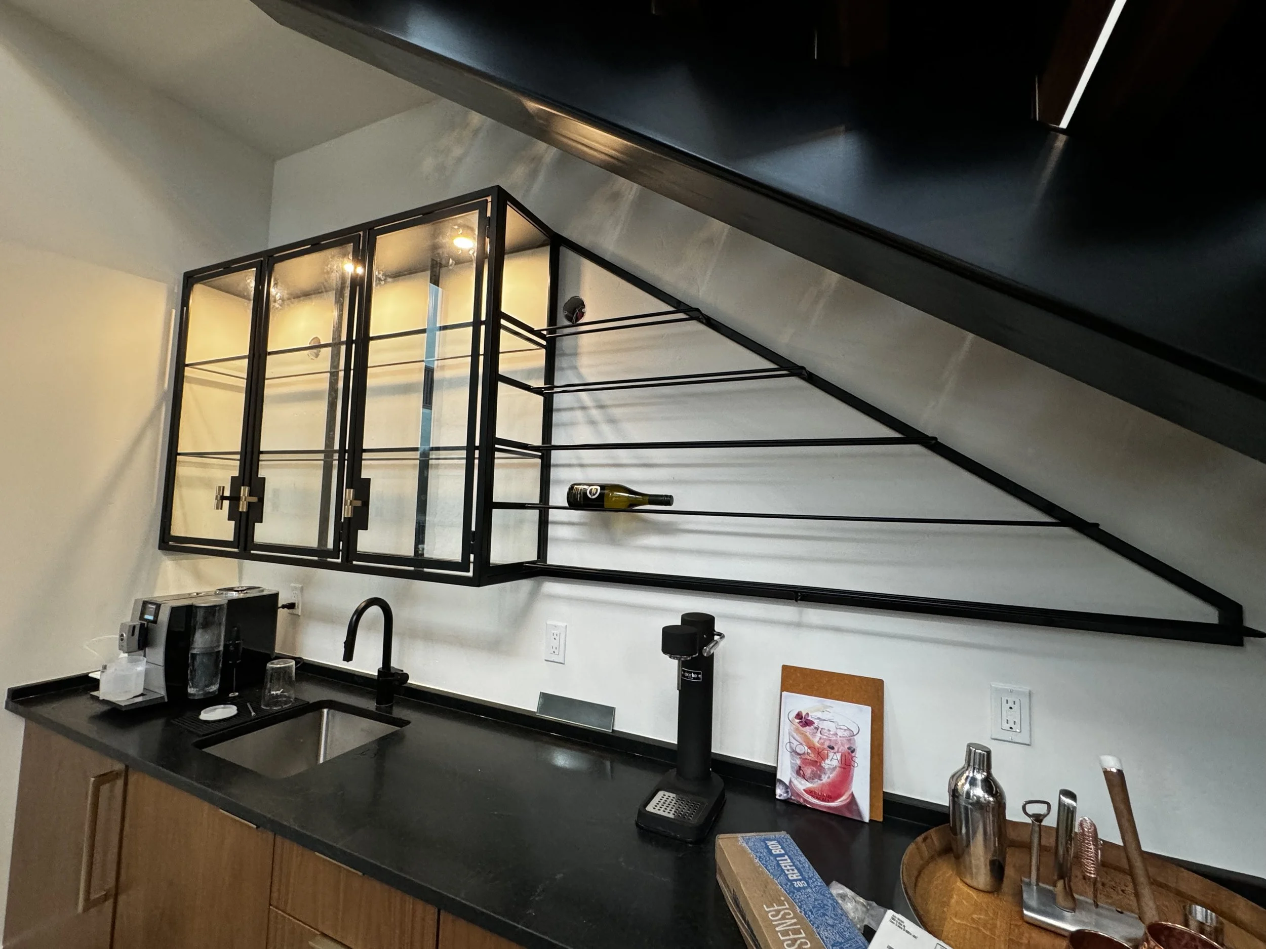 Modern kitchen with a black metal wall-mounted wine rack, a black countertop, a coffee machine, a glass, a water bottle, and a small corked bottle. The wall above the countertop has electrical outlets and a small framed menu or art piece. Wooden cabi