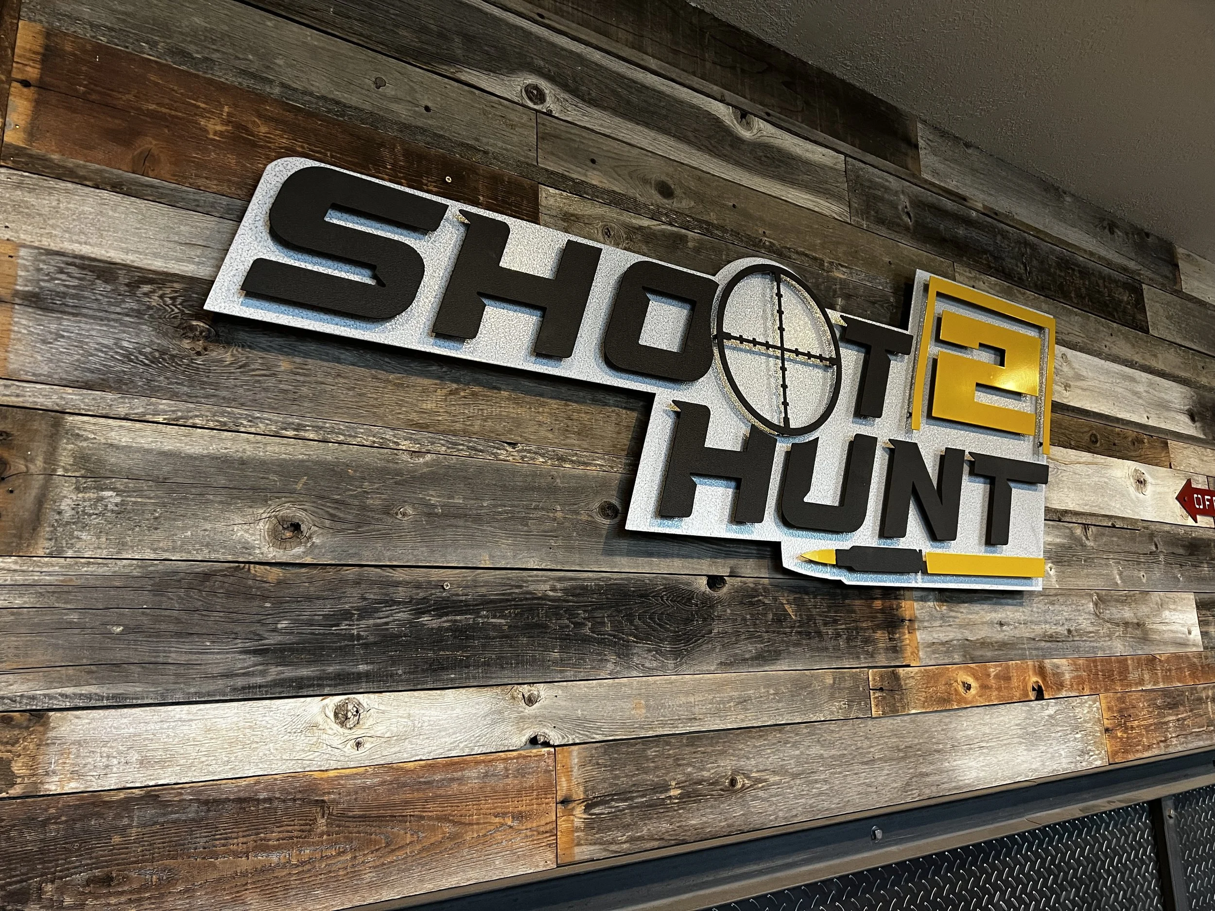 Sign for 'Shoot to Hunt' with a rifle scope crosshair graphic, mounted on a wooden wall.