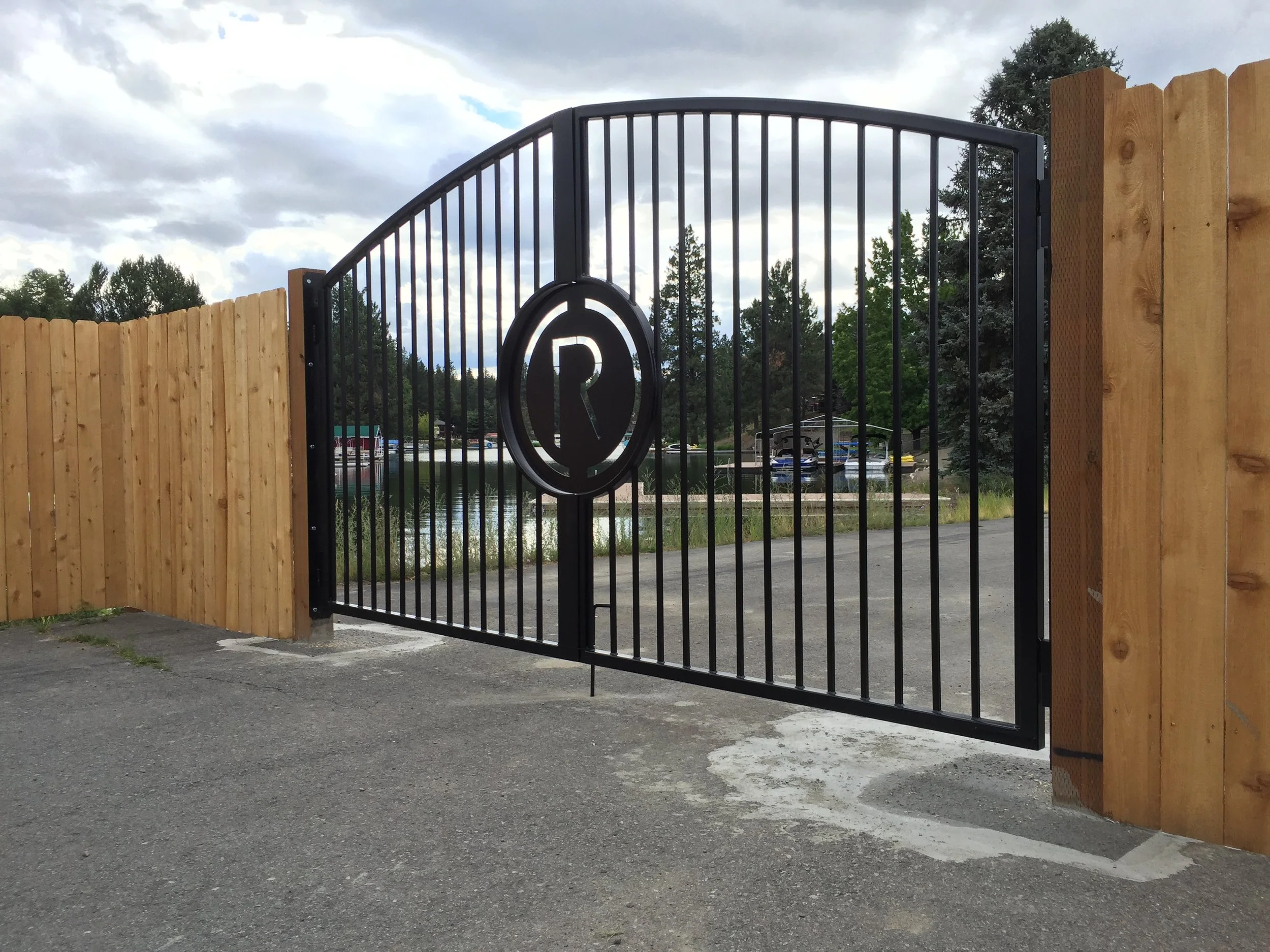 Black metal gate with a circular emblem featuring a stylized letter 'R', flanked by wooden fences, open to a view of boats docked by a pond, surrounded by trees under cloudy skies.