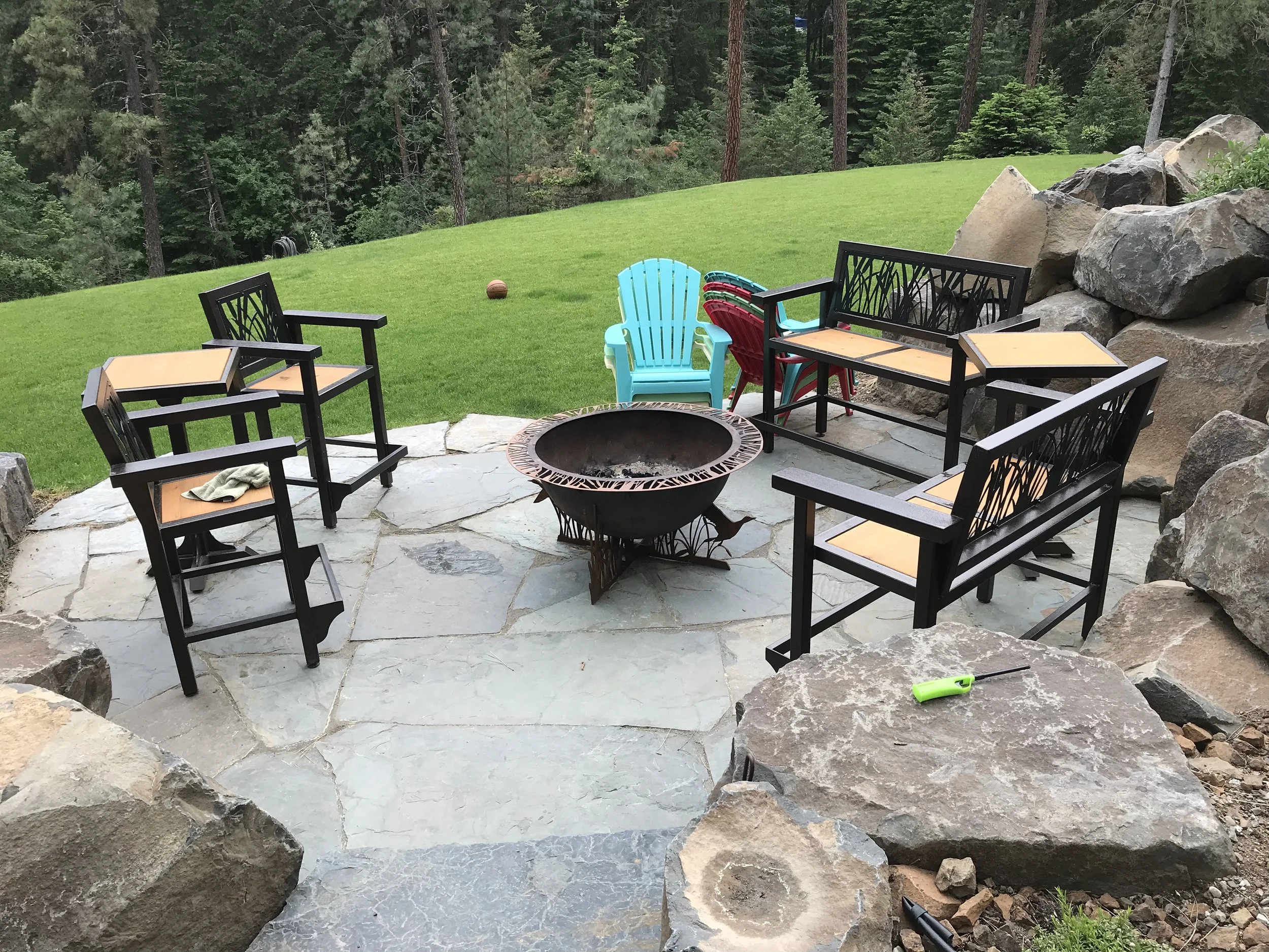 A backyard patio with black chairs around a fire pit, a blue Adirondack chair, and a lush green lawn with trees in the background.