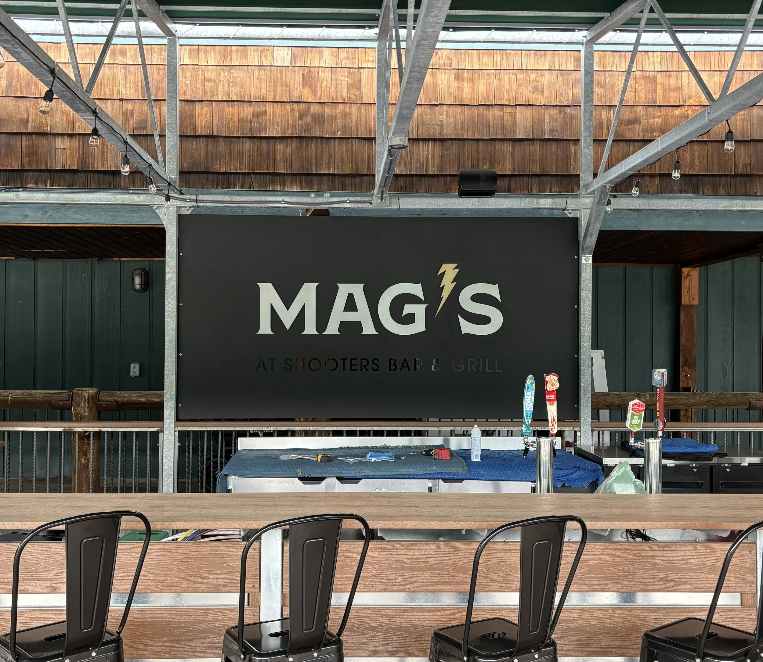 Interior of a restaurant or bar called MAG'S at Shooters Bar & Grill with a black sign, metal and wood decor, chairs, a bar counter, and beer taps.