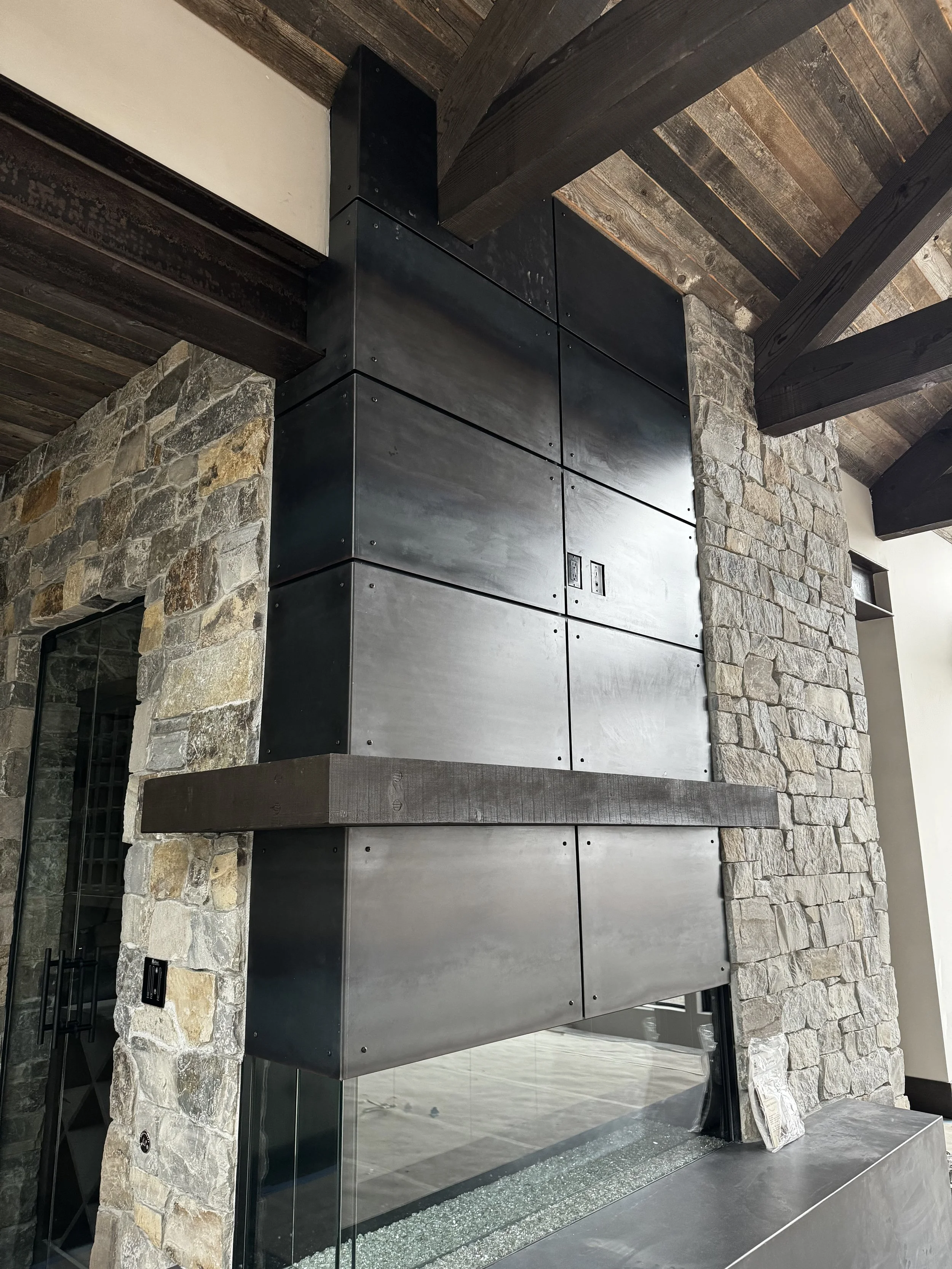 Interior view of a stone fireplace with a modern black metal and glass design, featuring a wooden mantle and dark wooden ceiling beams.