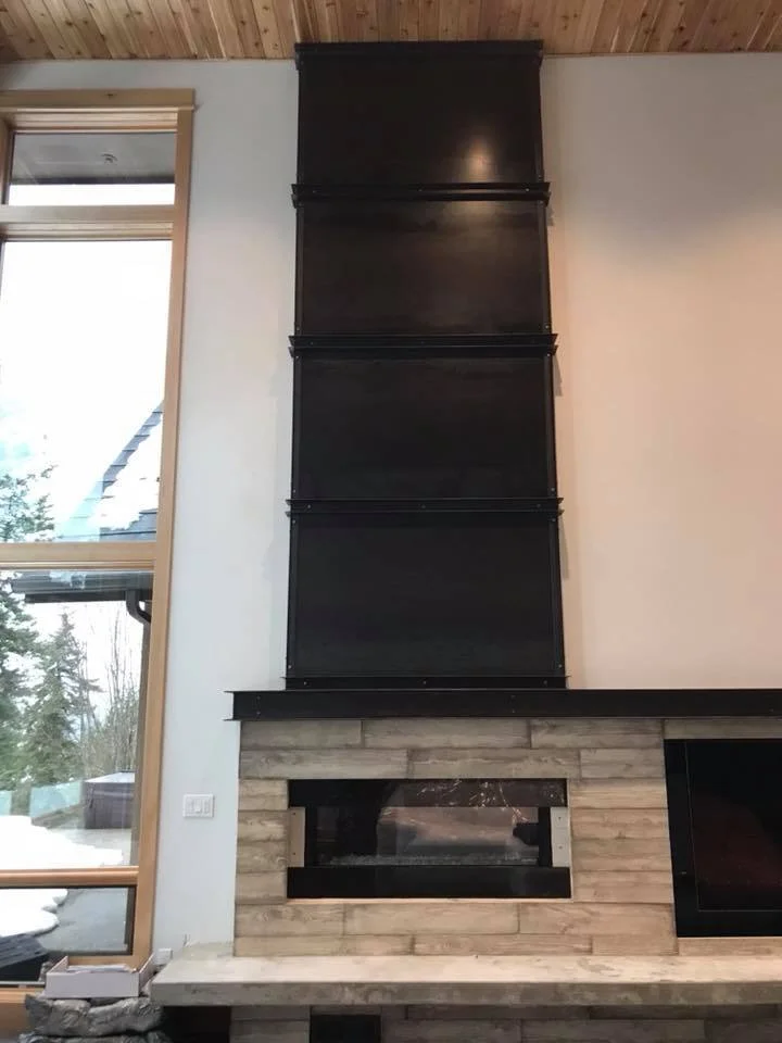Modern indoor fireplace with black metal chimney extending upward, surrounded by beige stone and wood. Large window to the left shows snowy outdoor scenery.