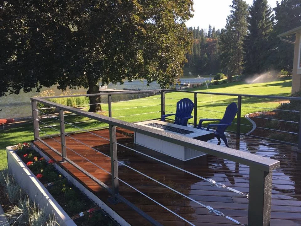 View of a wooden deck with two blue chairs, a white bench, and a railing overlooking a lake with trees and a grassy area in the background.