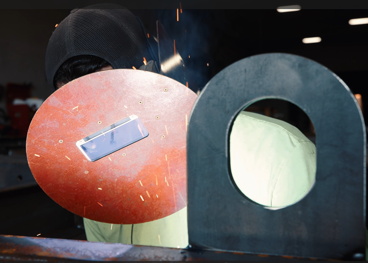 A person welding metal parts in a workshop, wearing a welding helmet and protective gear.