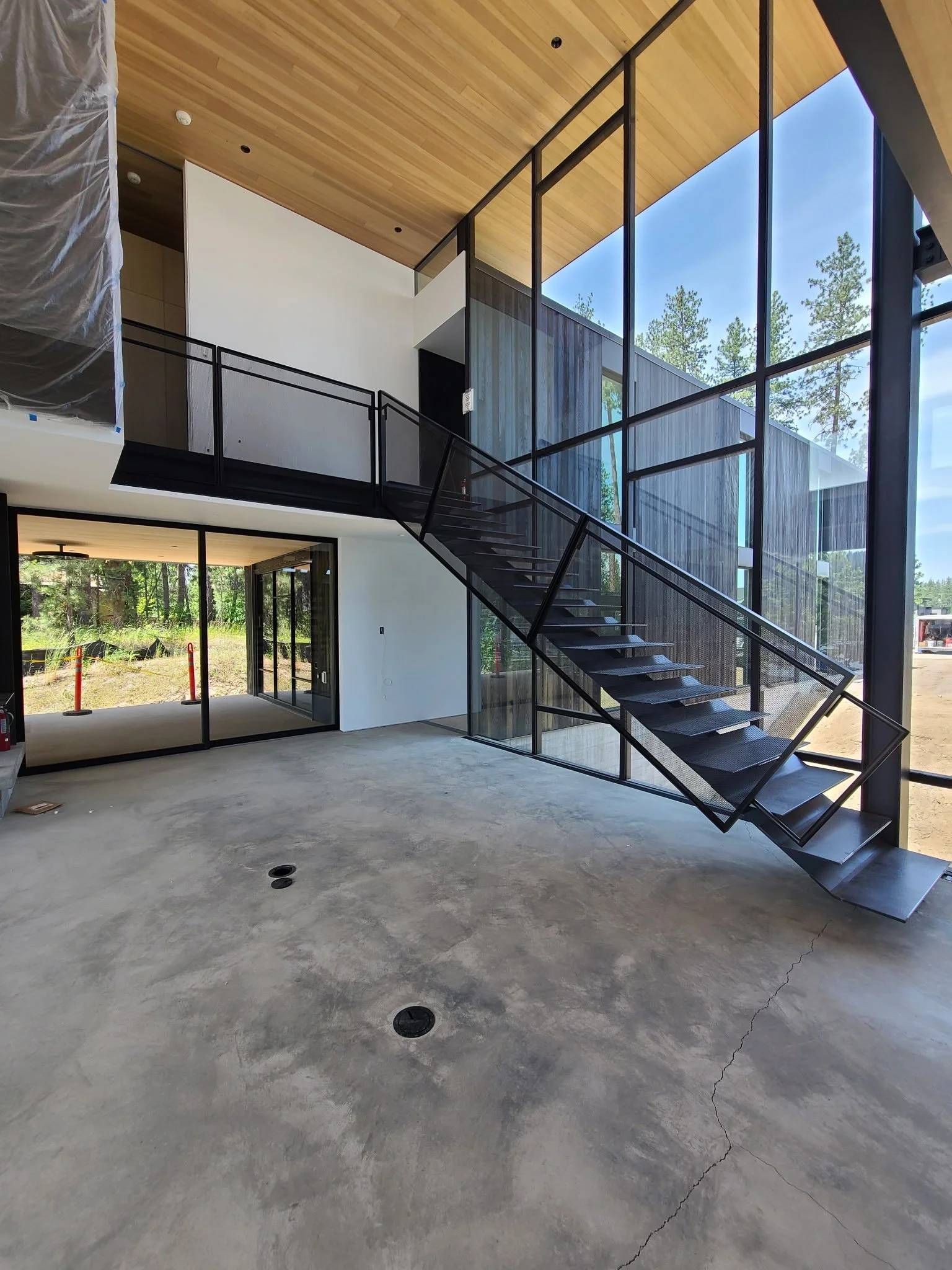 Interior of a modern house under construction with large floor-to-ceiling windows, black metal staircase, concrete floor, and wooden ceiling.