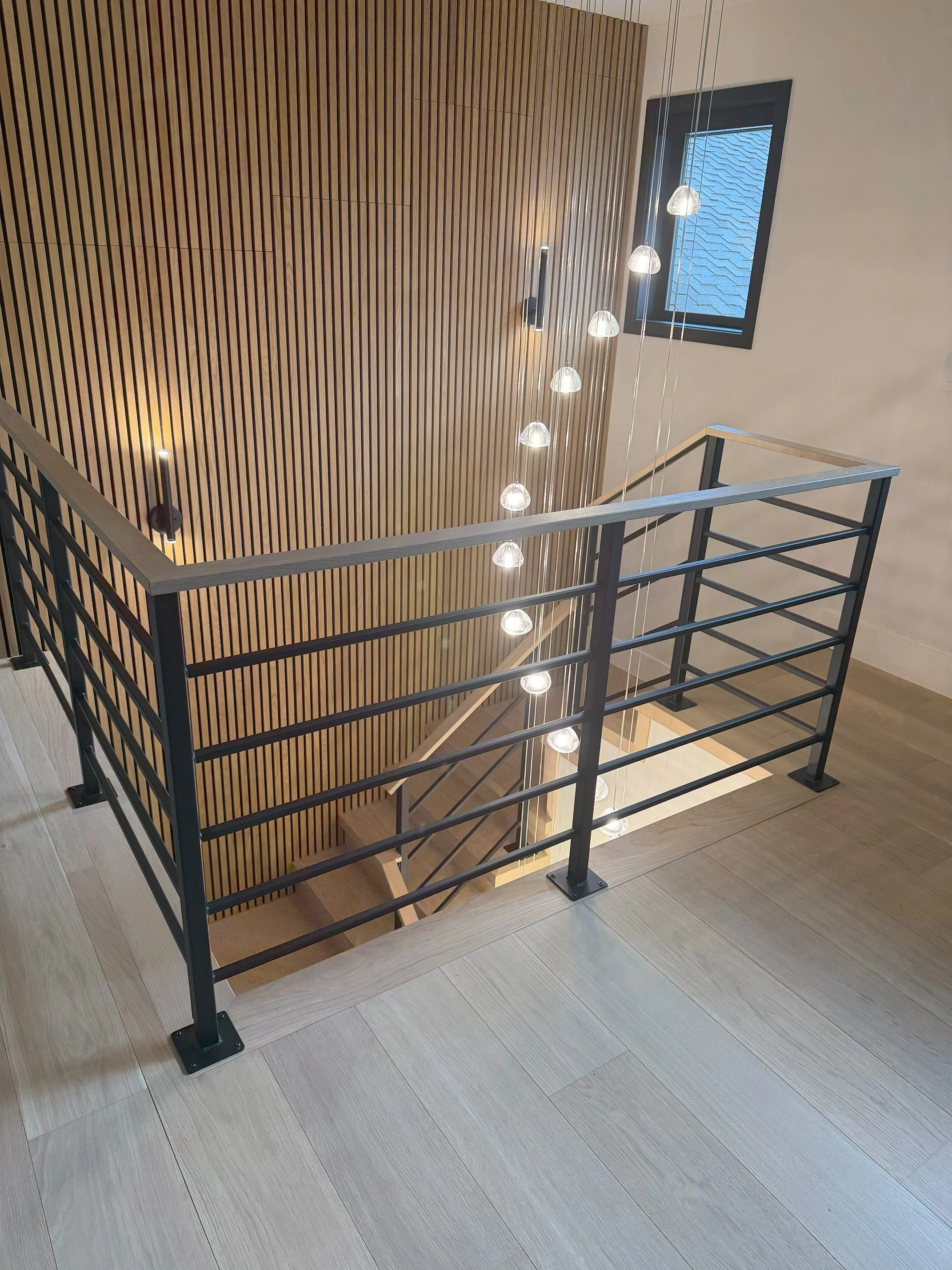 Indoor staircase with black metal railing, wooden steps, and a slatted wood wall. Modern hanging light fixtures and a small window with a blue roof exterior visible.