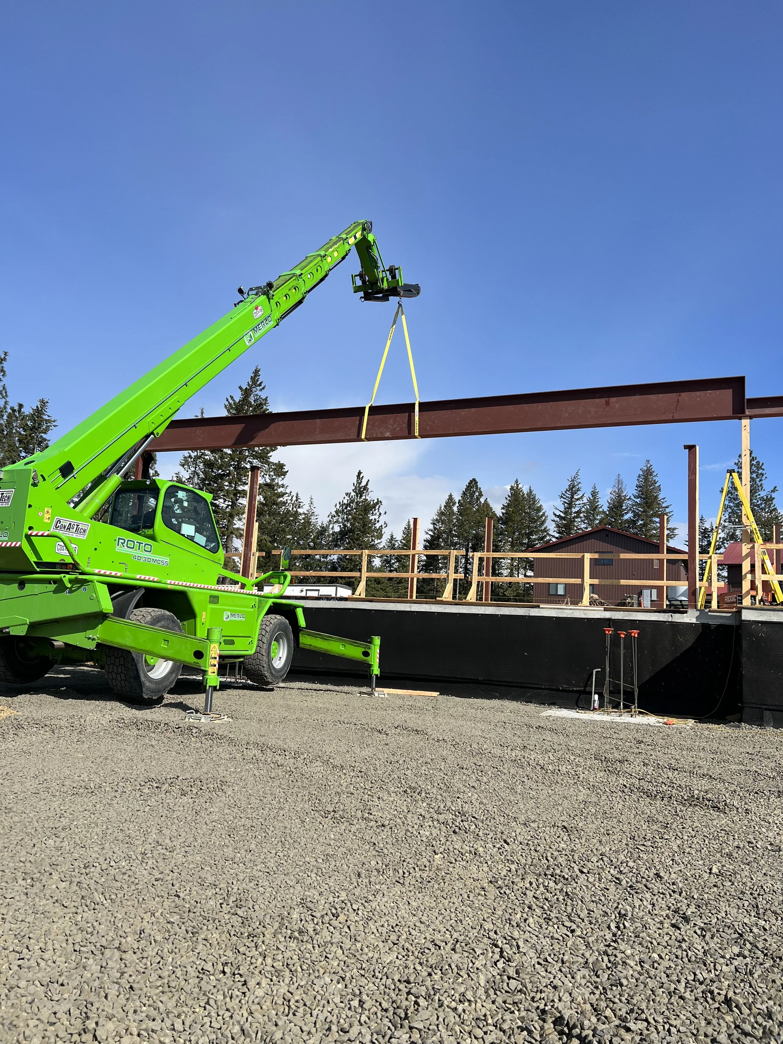 A bright green construction crane lifting a steel beam on a construction site with a gravel ground, framed by trees and a building with a red roof in the background.
