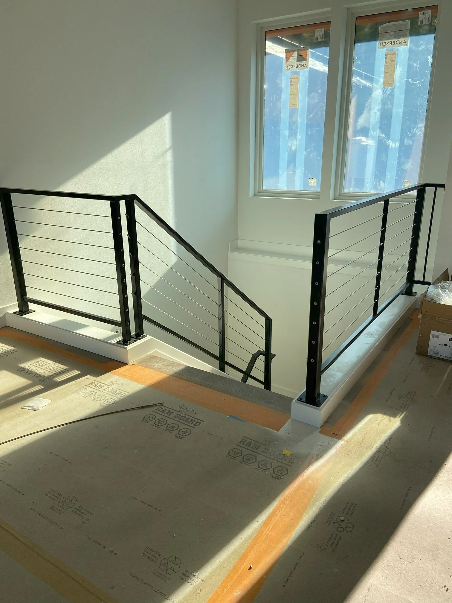 Interior view of a staircase with modern black metal railings and white base, next to a large window with sunlight streaming in, during construction or renovation.