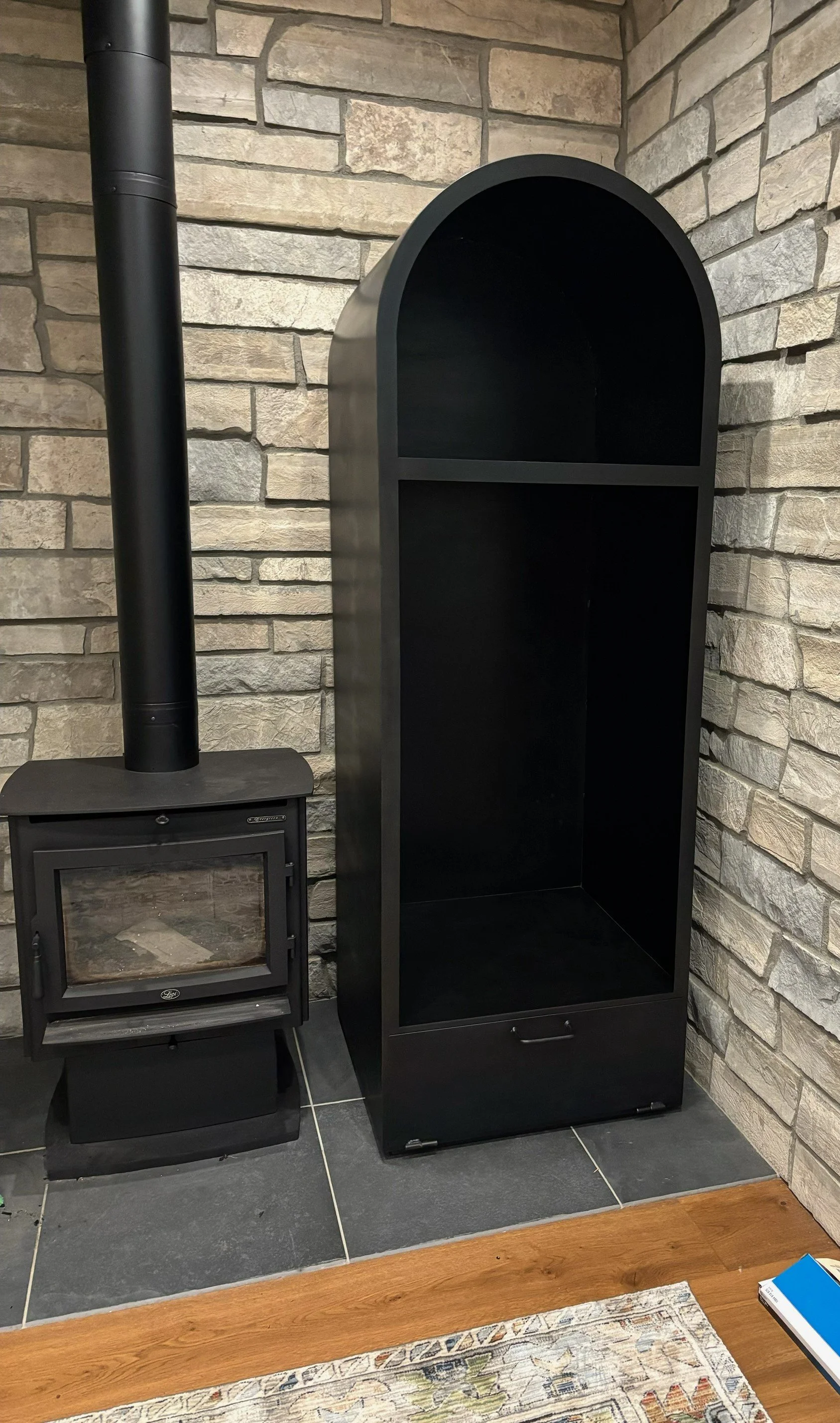 A black stove with a glass door and a chimney pipe, next to an empty black arched display cabinet against a stone wall.