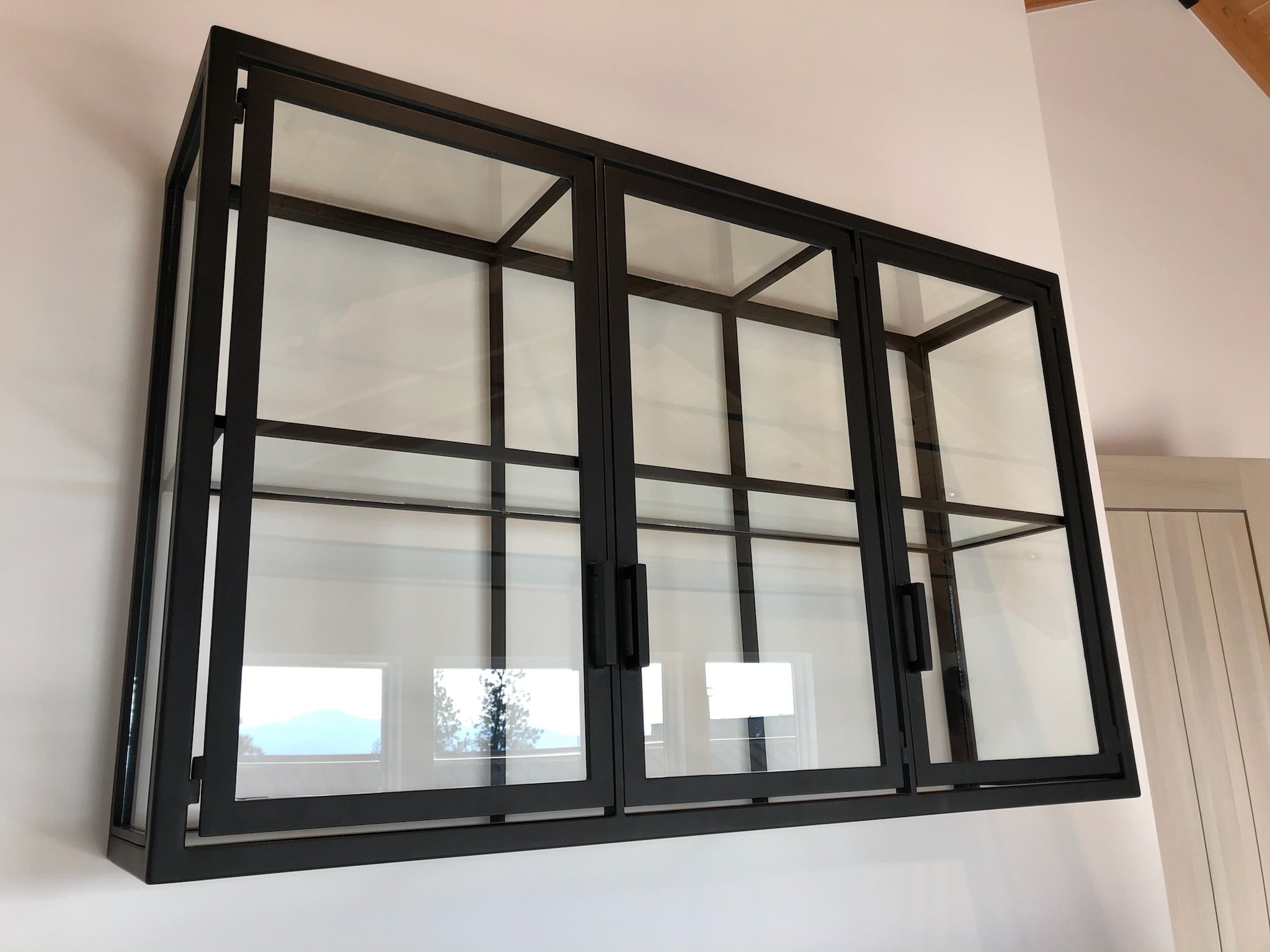 Black metal and glass wall-mounted display cabinet with sliding doors, empty inside, reflected windows and ceiling light visible.