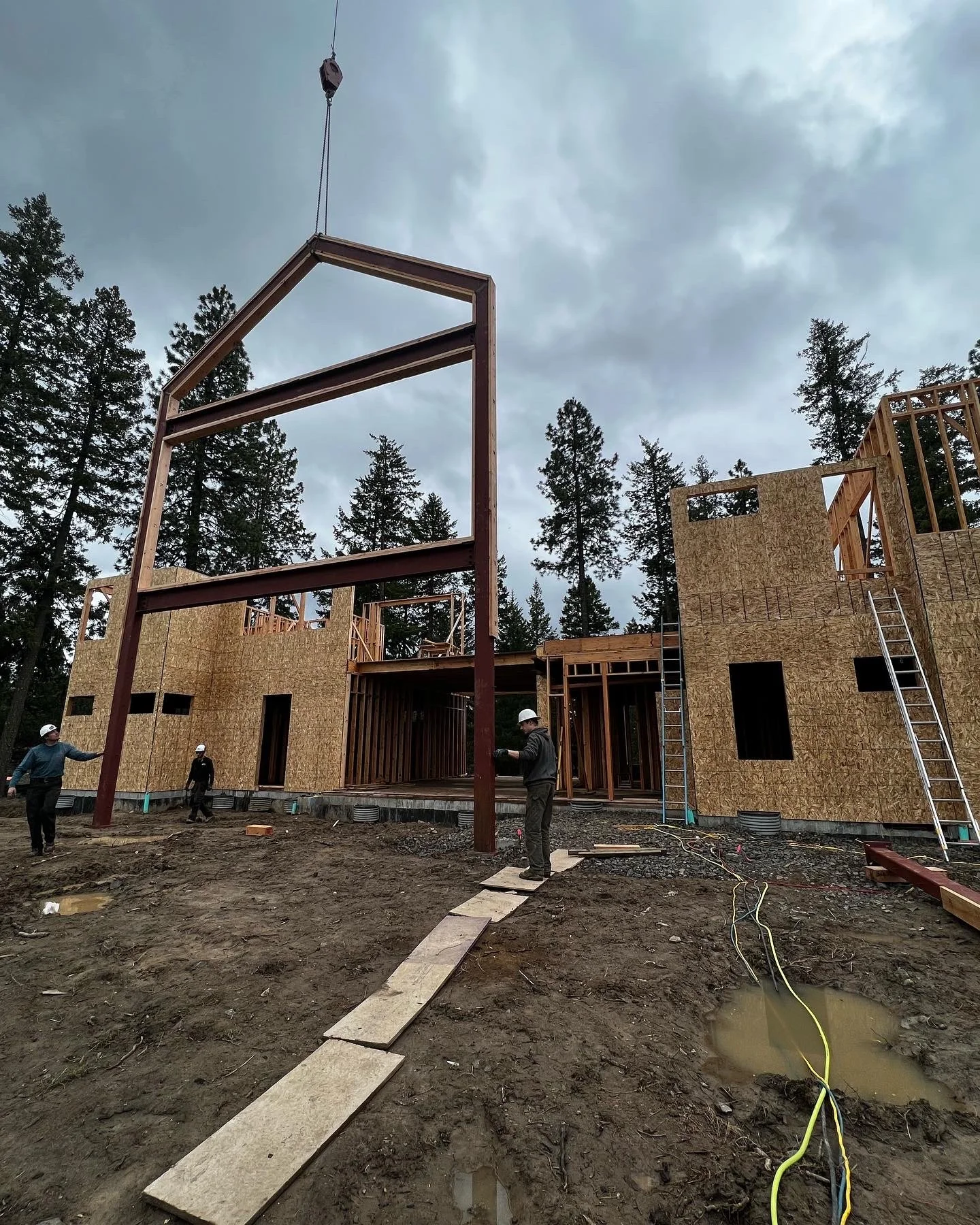Construction site with a partially built wooden house and large metal beams being installed, three workers in hard hats, and a cloudy sky.