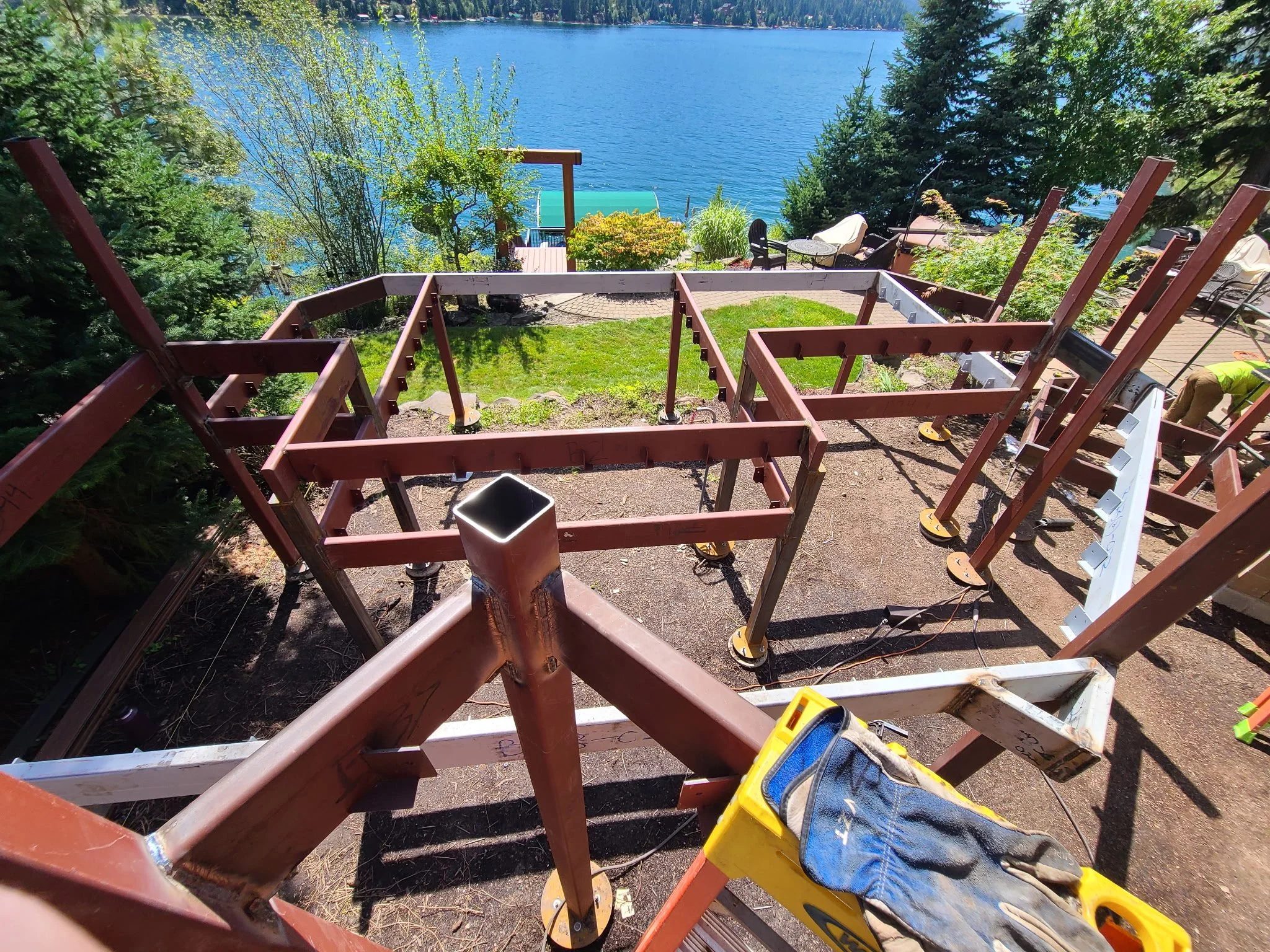 Construction of a deck or patio over a hillside with a lake view, showing building frames, tools, and workers.