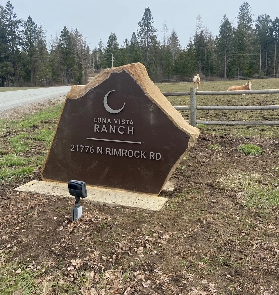 A large stone sign at Luna Vista Ranch with a crescent moon symbol, showing the address 21776 N Rimrock Rd, situated near a rural road and a fenced pasture with two horses, trees in the background, and a small light in front of the sign.