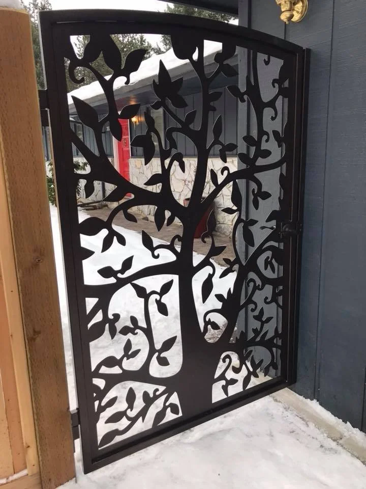 Decorative black metal gate with a tree and bird design, installed at an outdoor entrance with snow and a stone wall in the background.