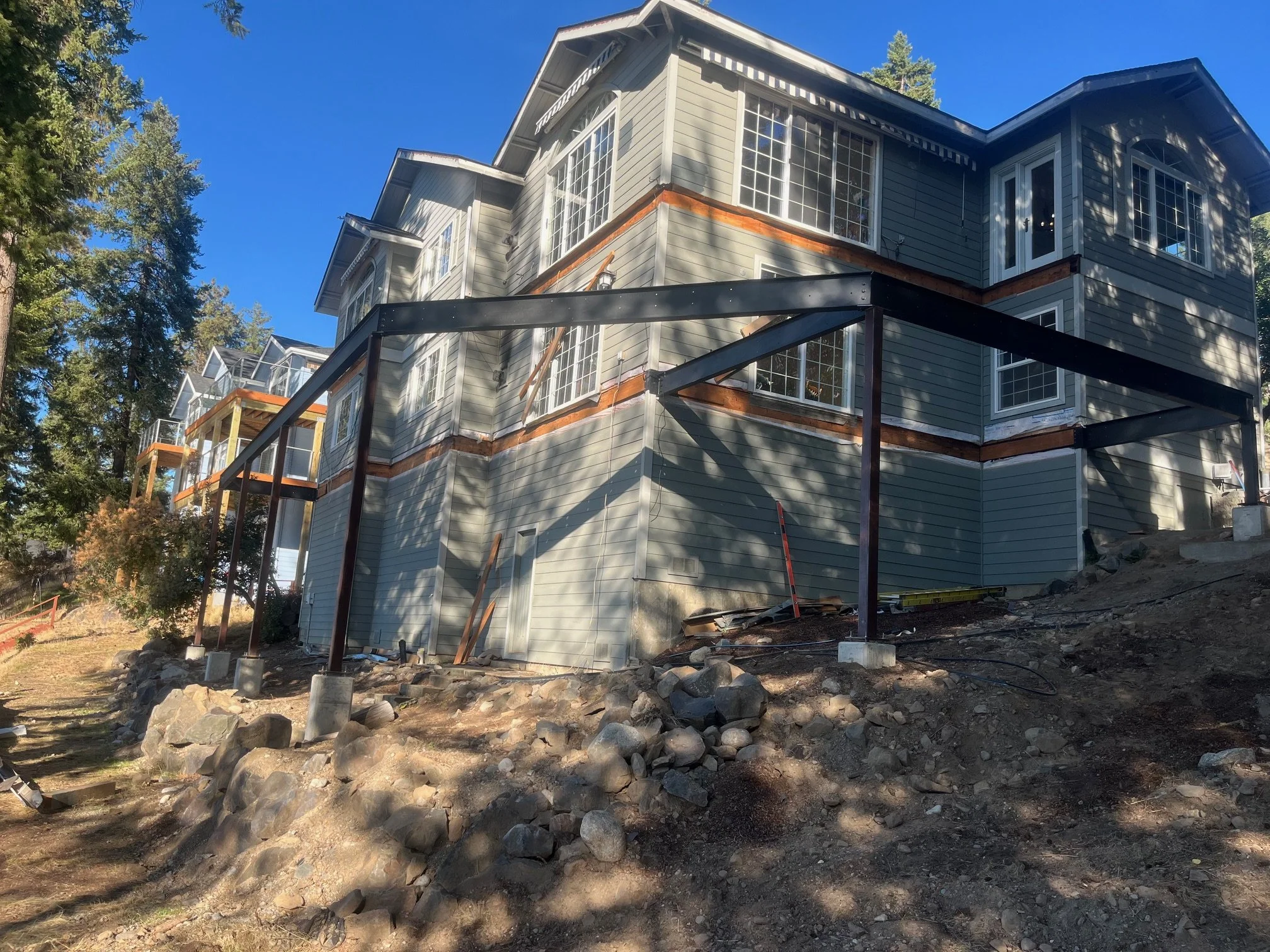 A multi-story house under construction with a dark metal frame on a sloped lot, surrounded by trees and rocks, with multiple large windows and blue siding.
